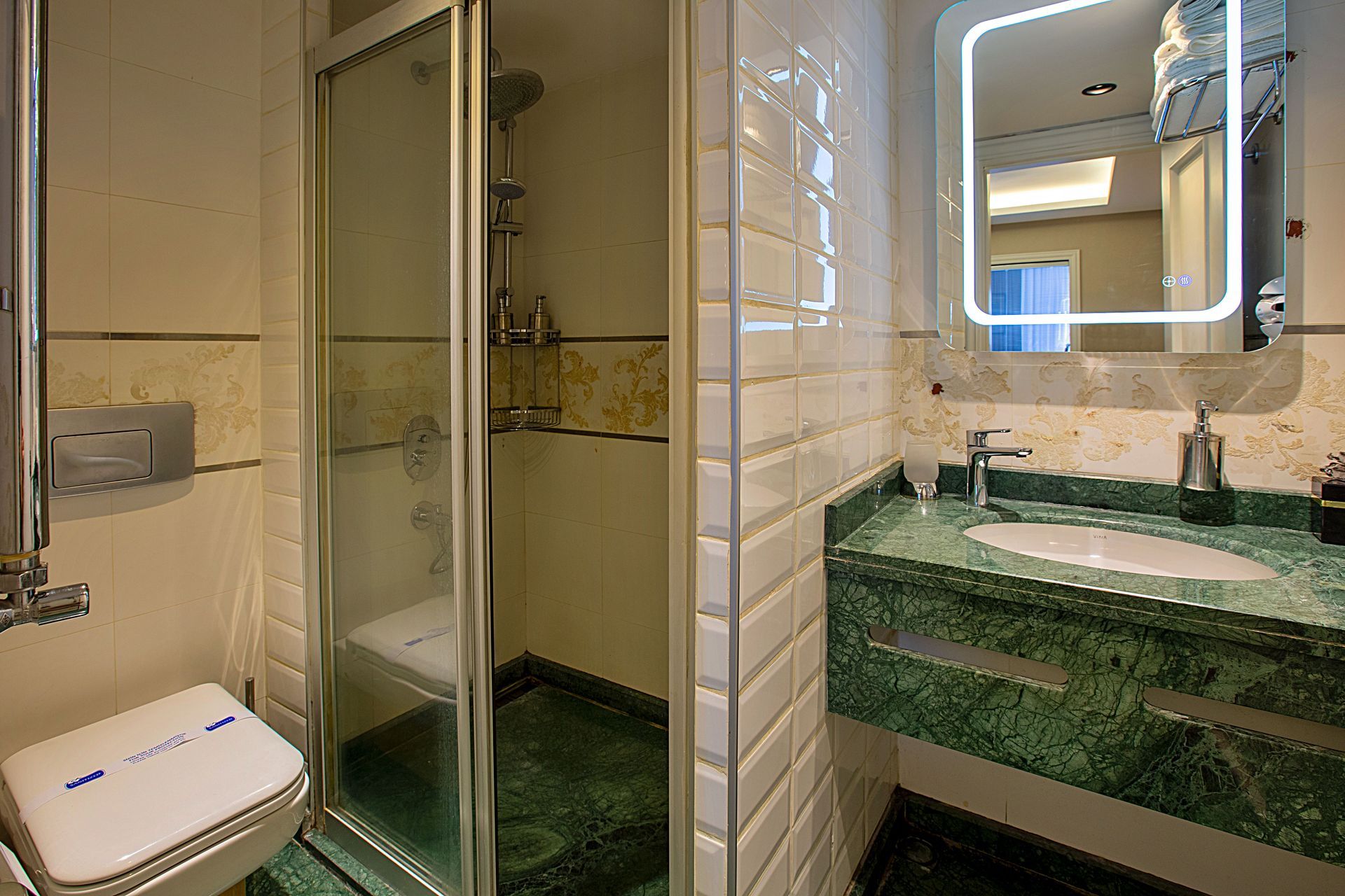 Compact bathroom with glass shower, toilet, and green marble sink vanity with lit mirror