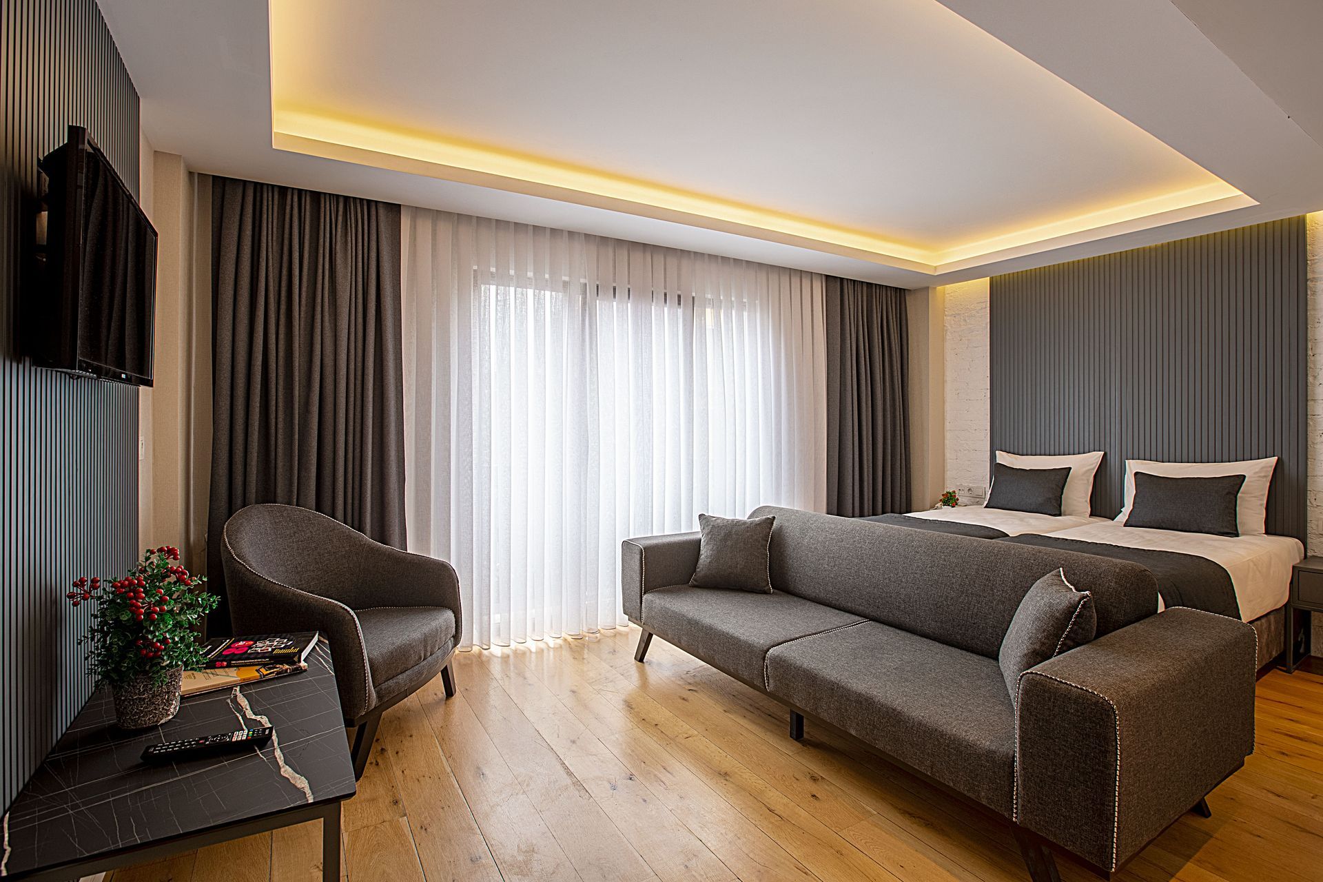Gri kanepe, sandalye ve yatağın bulunduğu, açık renkli ahşap zeminli ve tavandan tabana pencereli otel odası.