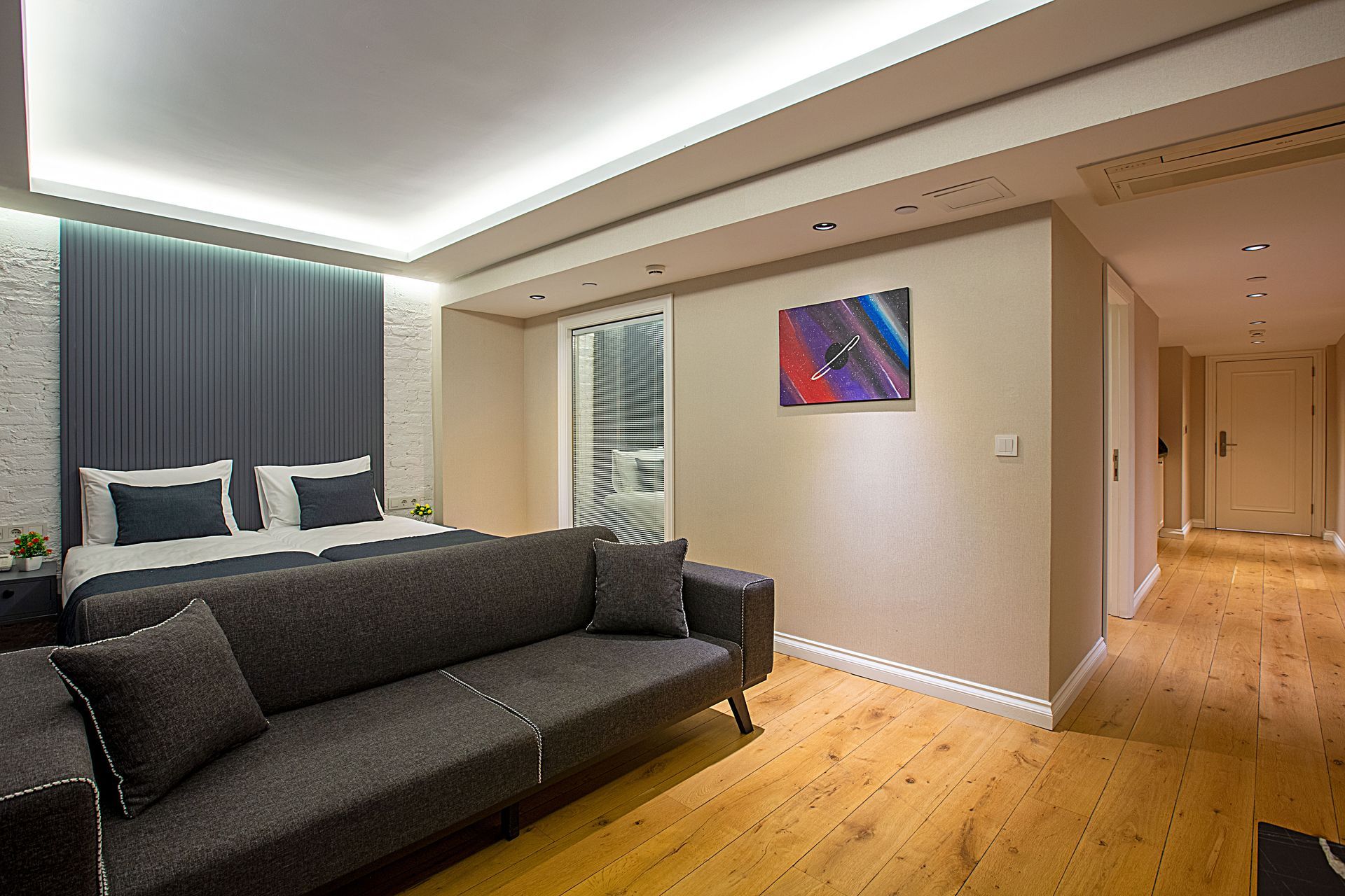 Modern bedroom with dark sofa, bed, wood floors, and hallway lighting