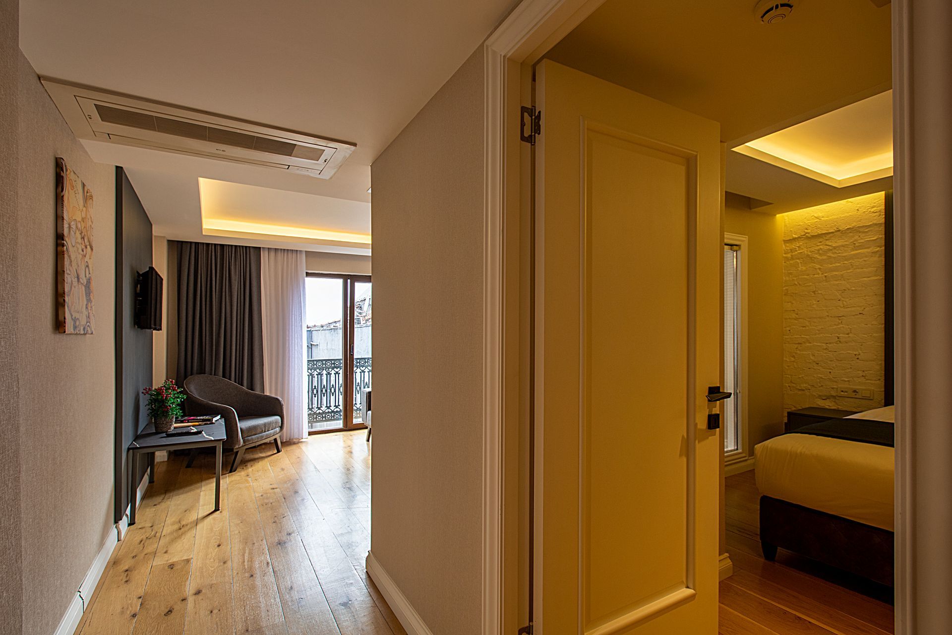 Bright hallway leading to a bedroom with an open door, wood floors, and warm yellow lighting.