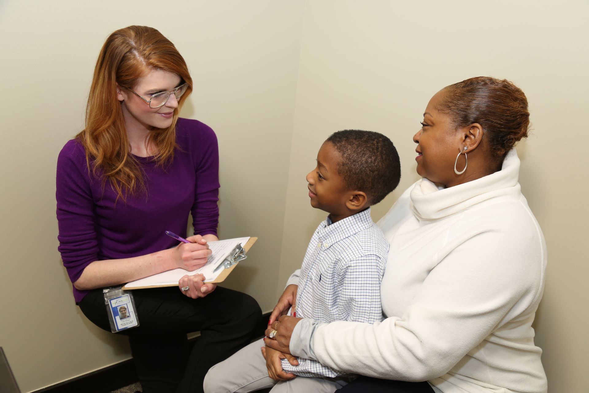 Woman interviewing a boy and his mother in an office. The woman is wearing purple, and writing notes.