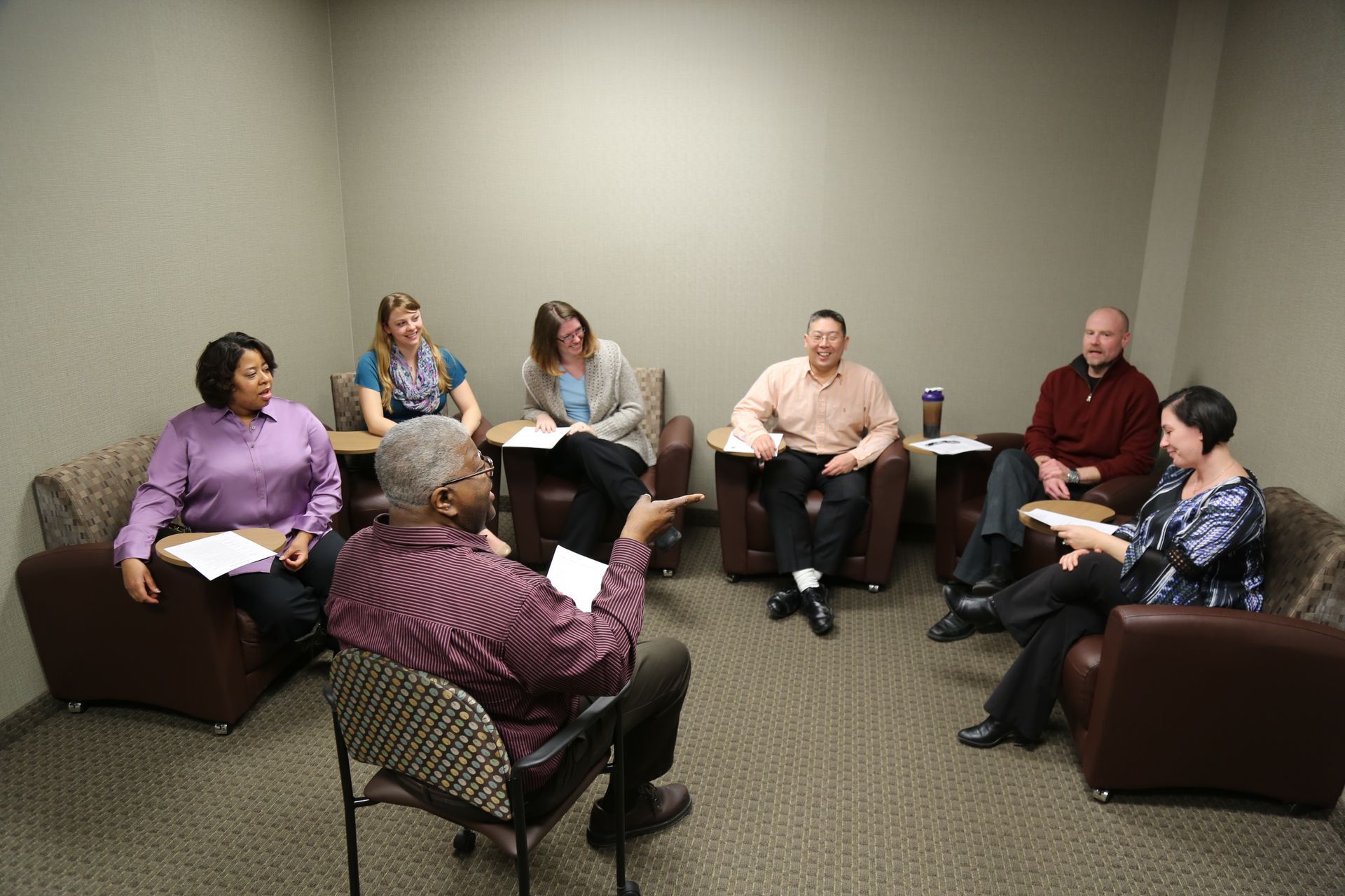 Group of diverse people in armchairs in a circle, listening to a speaker. Beige walls, neutral carpet.