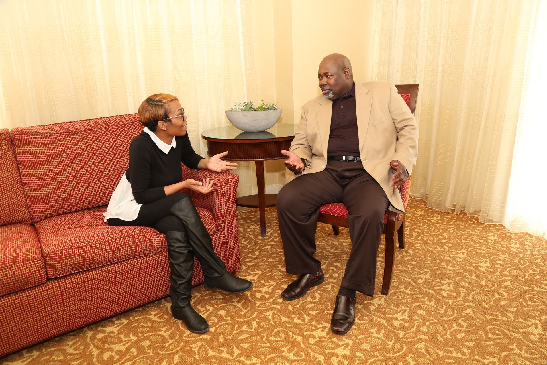 Two people talking on a red couch in a small living room with curtains and a round table.