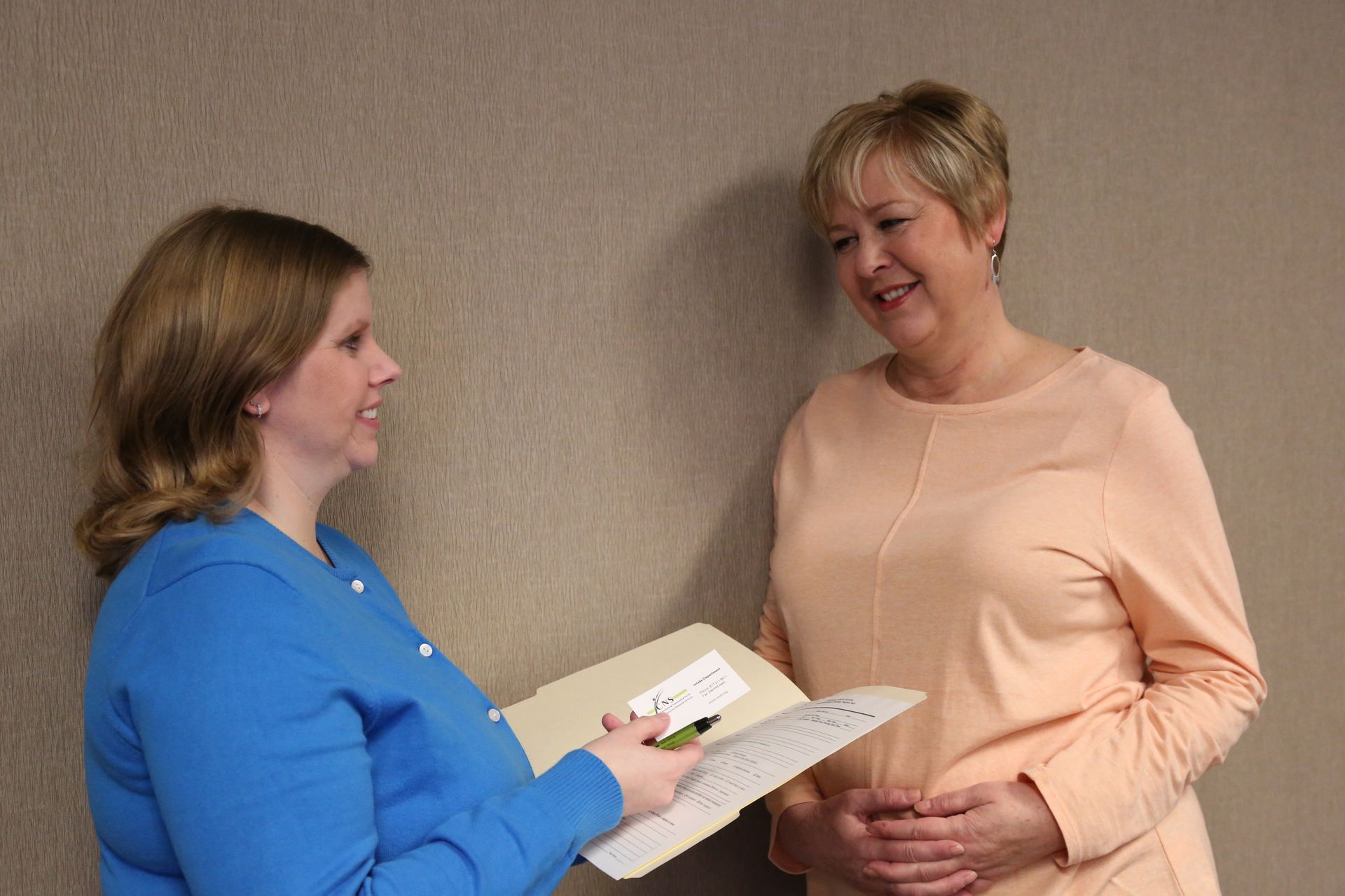 Two women talking indoors, one holding a folder and paper while the other smiles in a peach top.