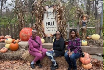 Three women are sitting on a hay bale in front of a sign that says `` fall ''.