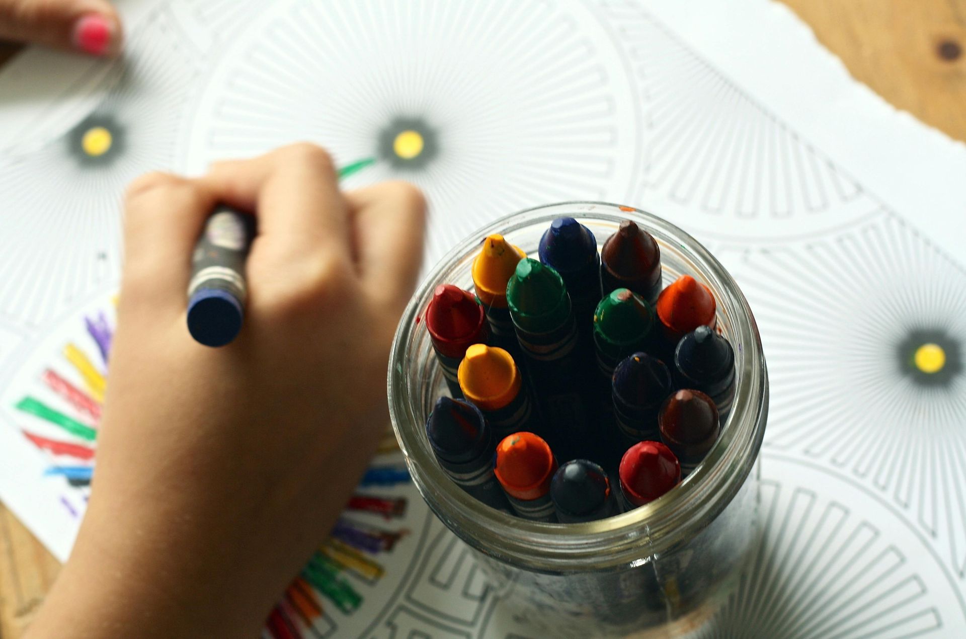 Child coloring a detailed mandala design with crayons, jar of crayons nearby.