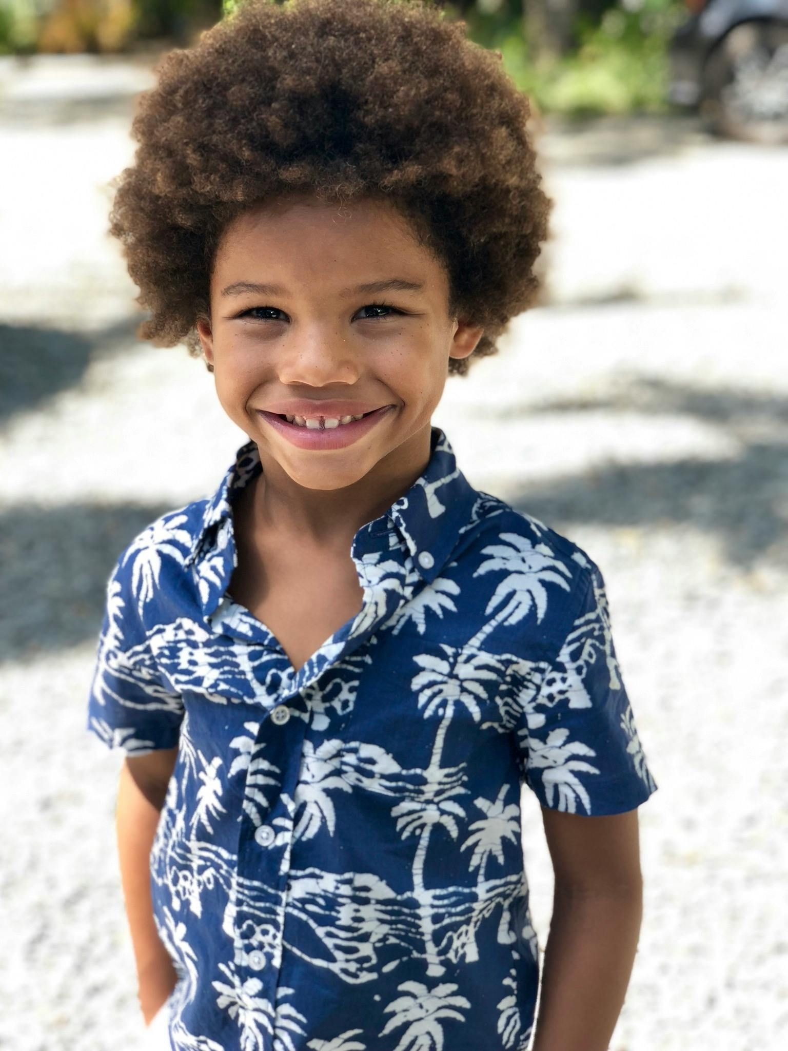 Young boy with curly brown hair smiles, wearing a blue and white palm tree print shirt.
