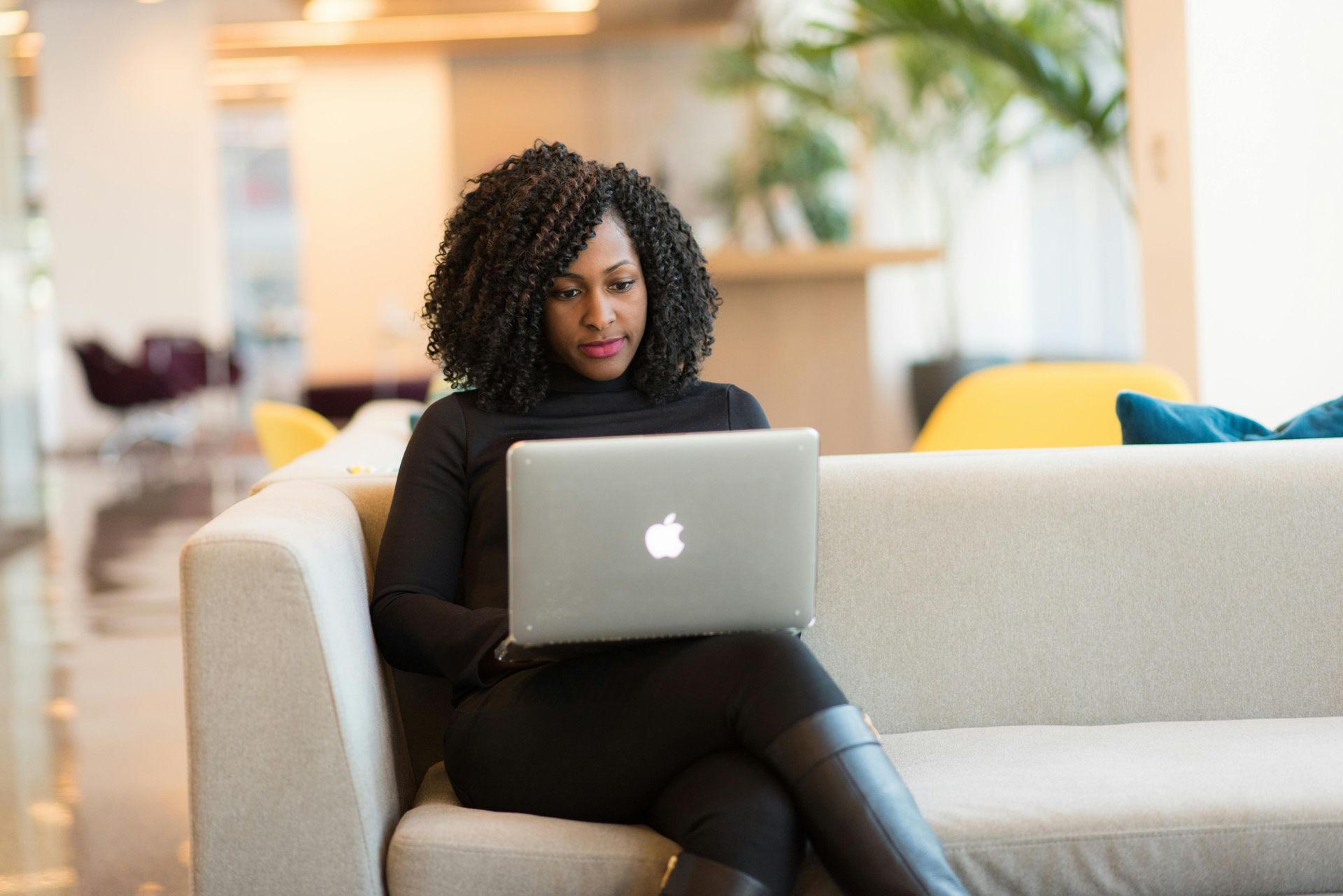 Woman with curly hair works on a laptop while sitting on a light-colored couch in a modern office.