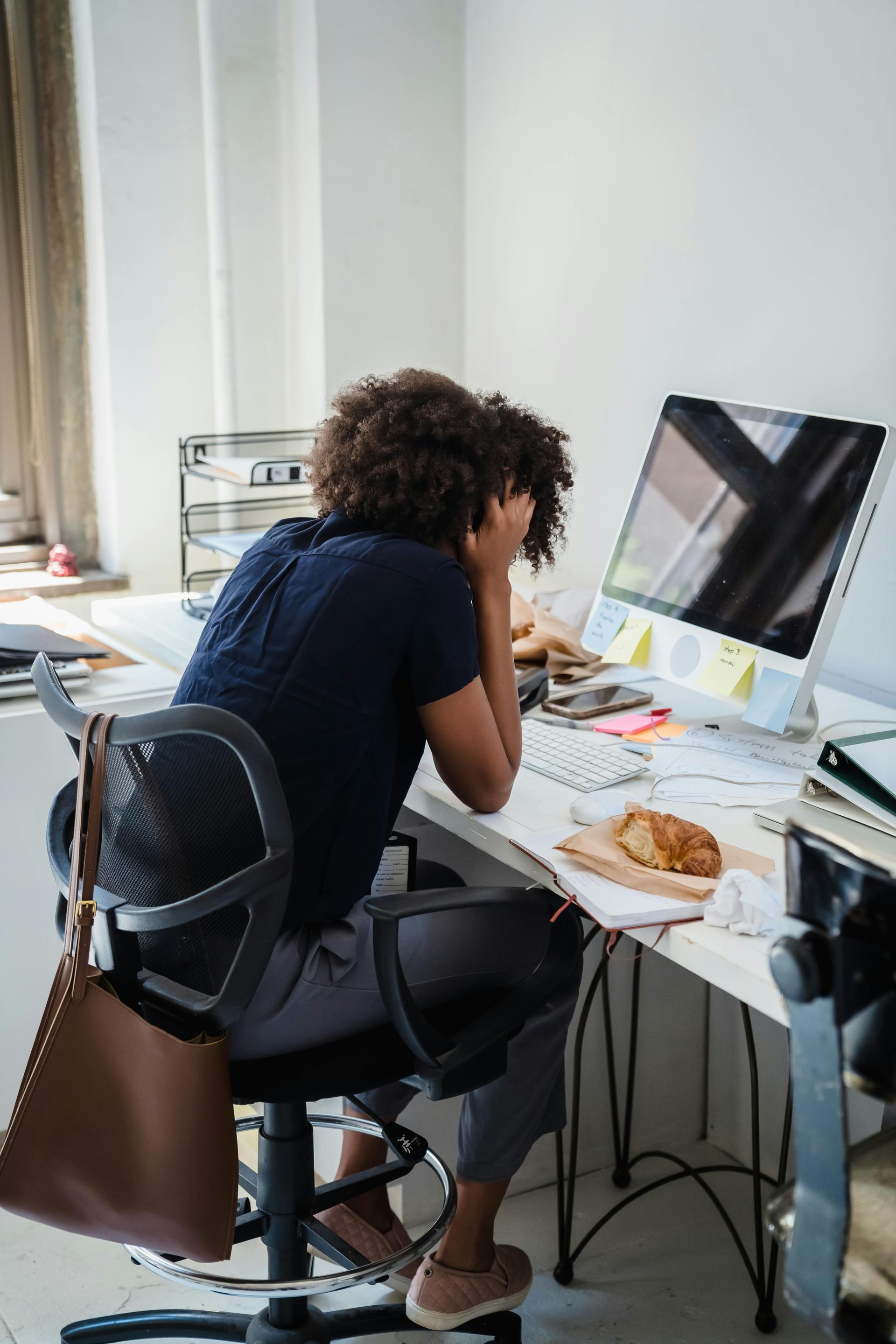 Person sitting at a messy desk, head in hands, appears stressed. A computer, papers, and food are visible.