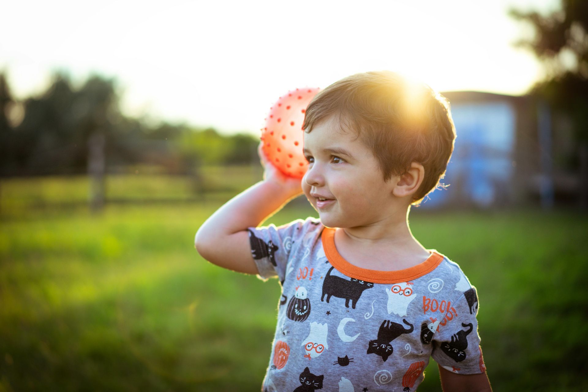 Child in a patterned shirt holding a red ball outdoors at sunset with a blurred grassy background