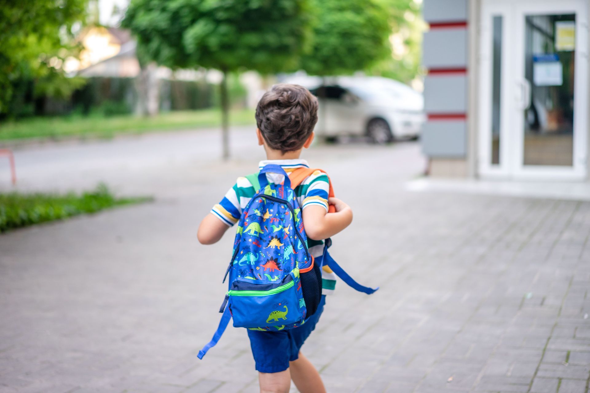 Child with blue backpack walking on a sidewalk toward a building entrance