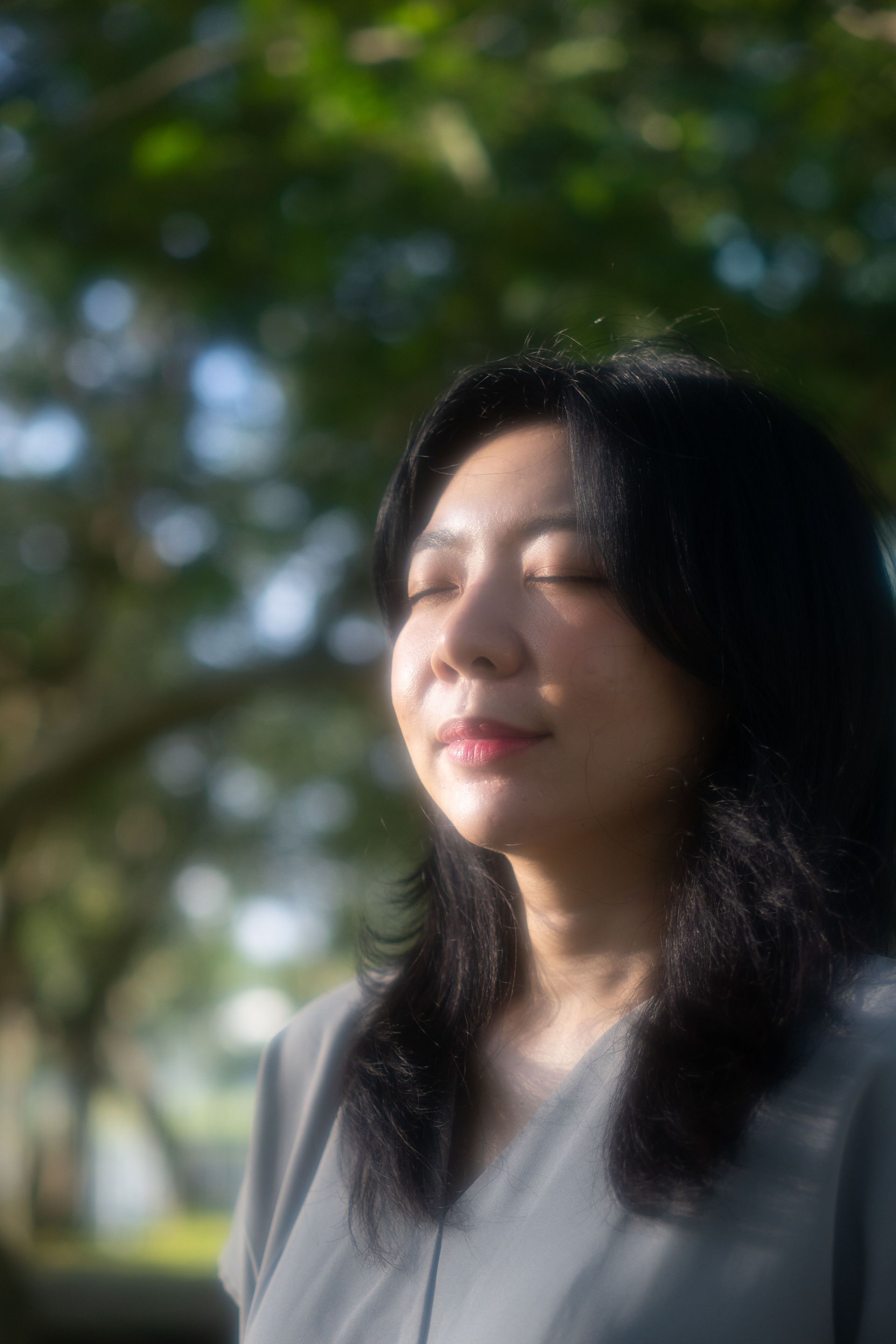 Person with eyes closed, standing outdoors in sunlight with trees softly blurred behind.