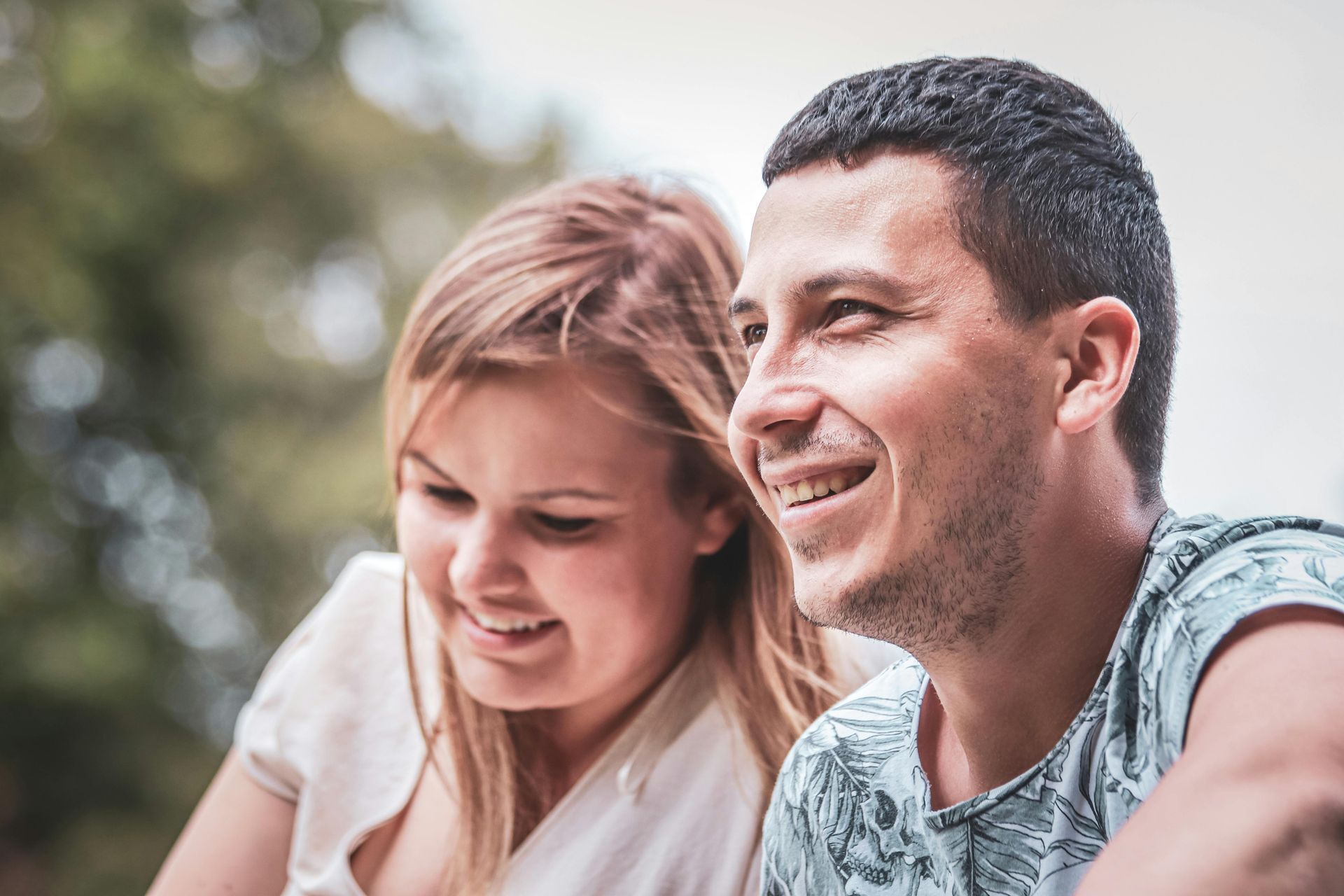 Two smiling people outdoors, one in a white top and one in a patterned shirt, with a blurred background