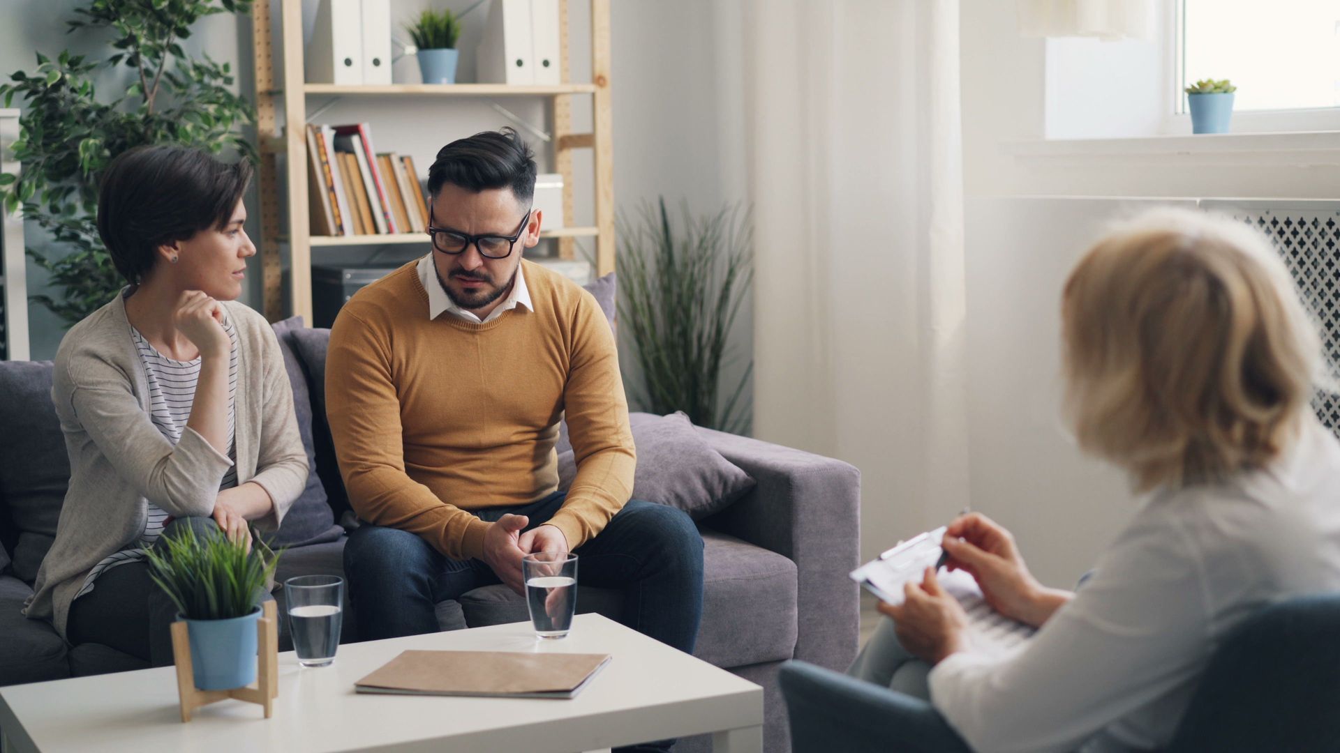 Three people in a bright office meeting, seated on a couch and chair around a coffee table.
