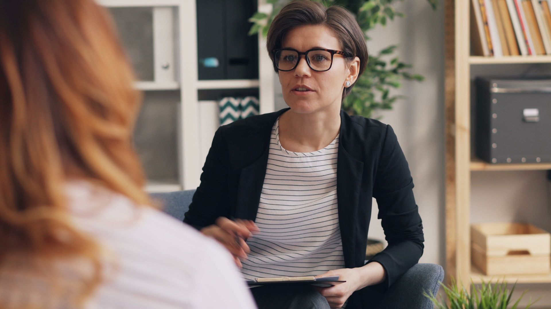 Woman in black cardigan speaking during a counseling session in an office, holding a clipboard.