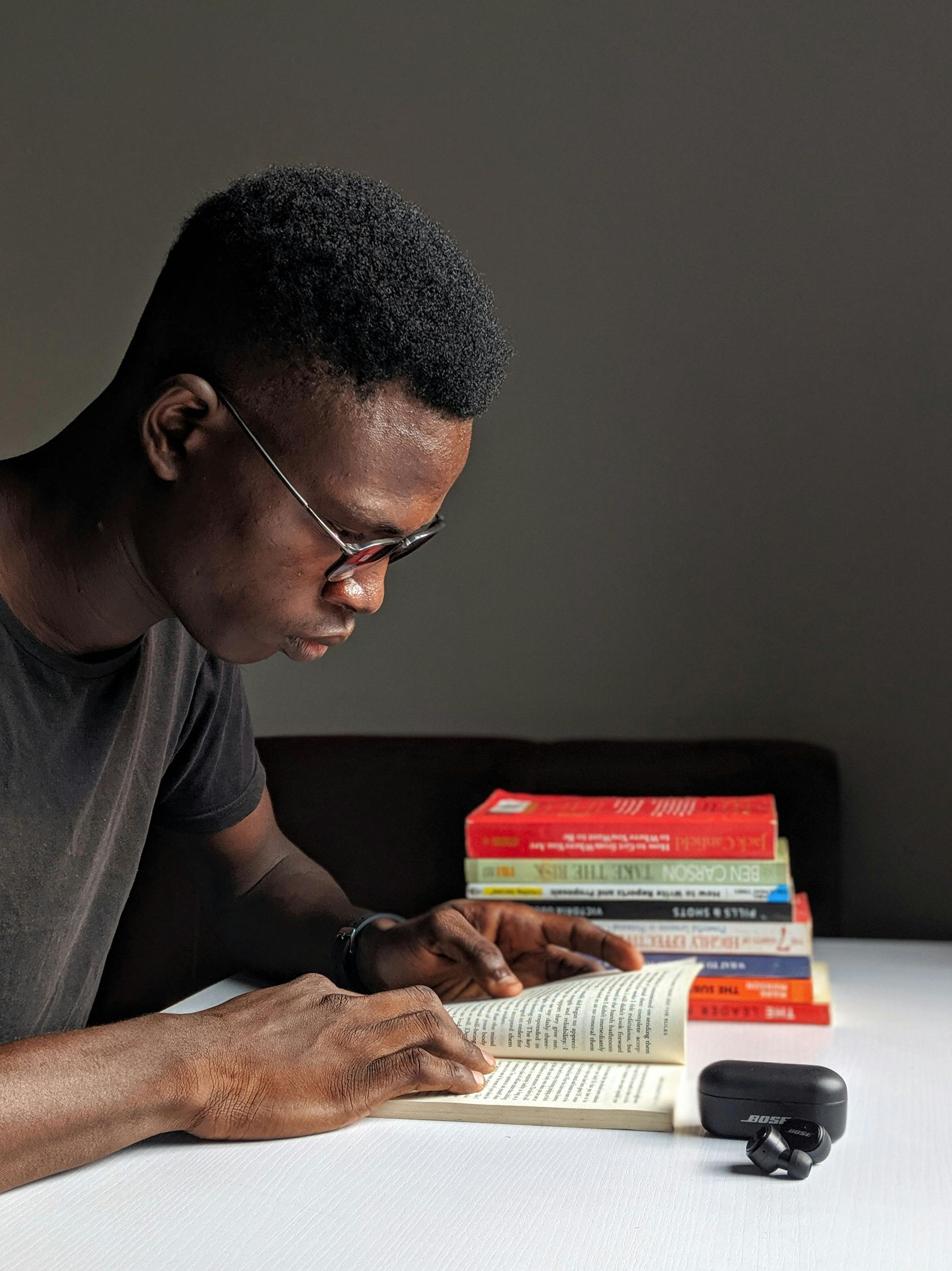 A person wearing glasses reads a book, with a stack of books behind them on a white surface.