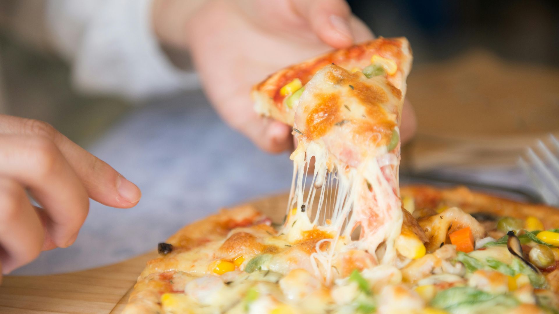 Hand lifting a cheesy pizza slice, cheese stretching, over the rest of the pizza on a wooden board.