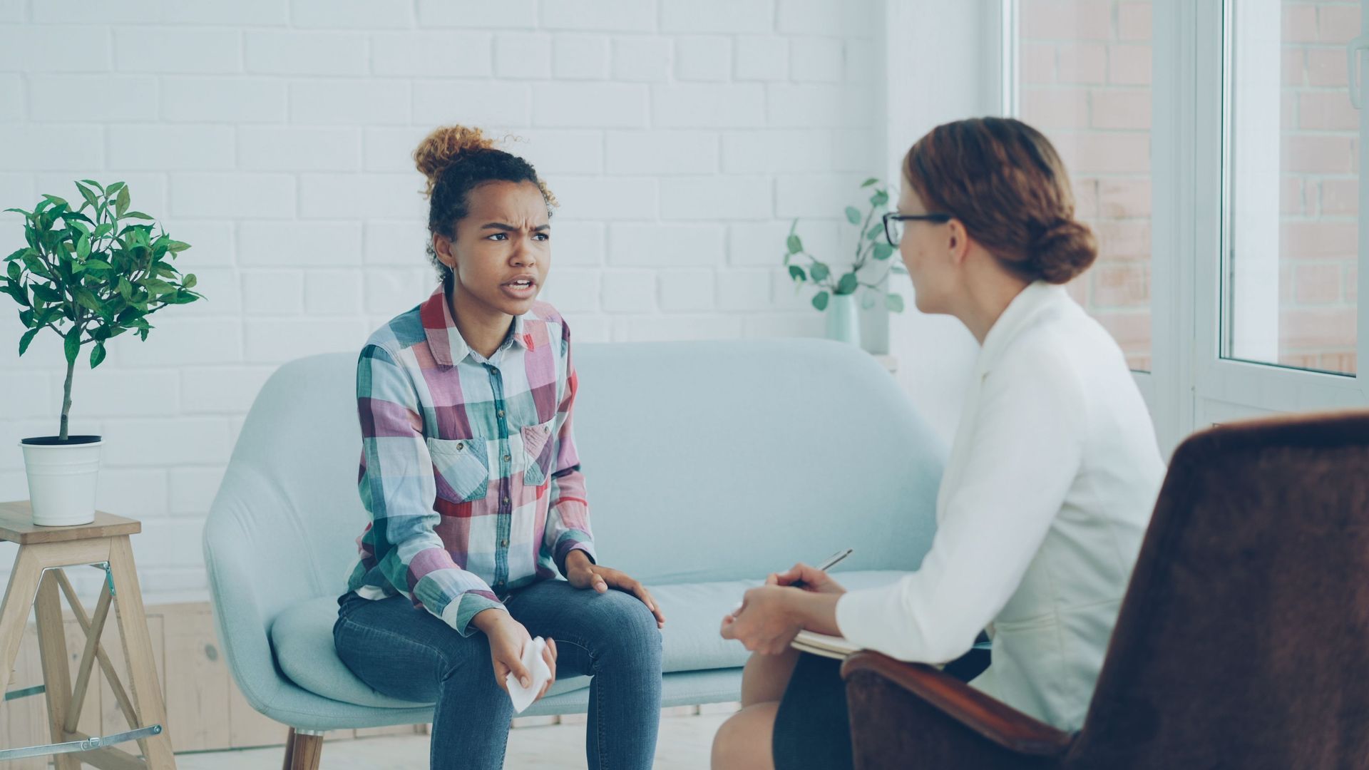 Two people in a counseling session, seated and talking in a bright office with a potted plant.