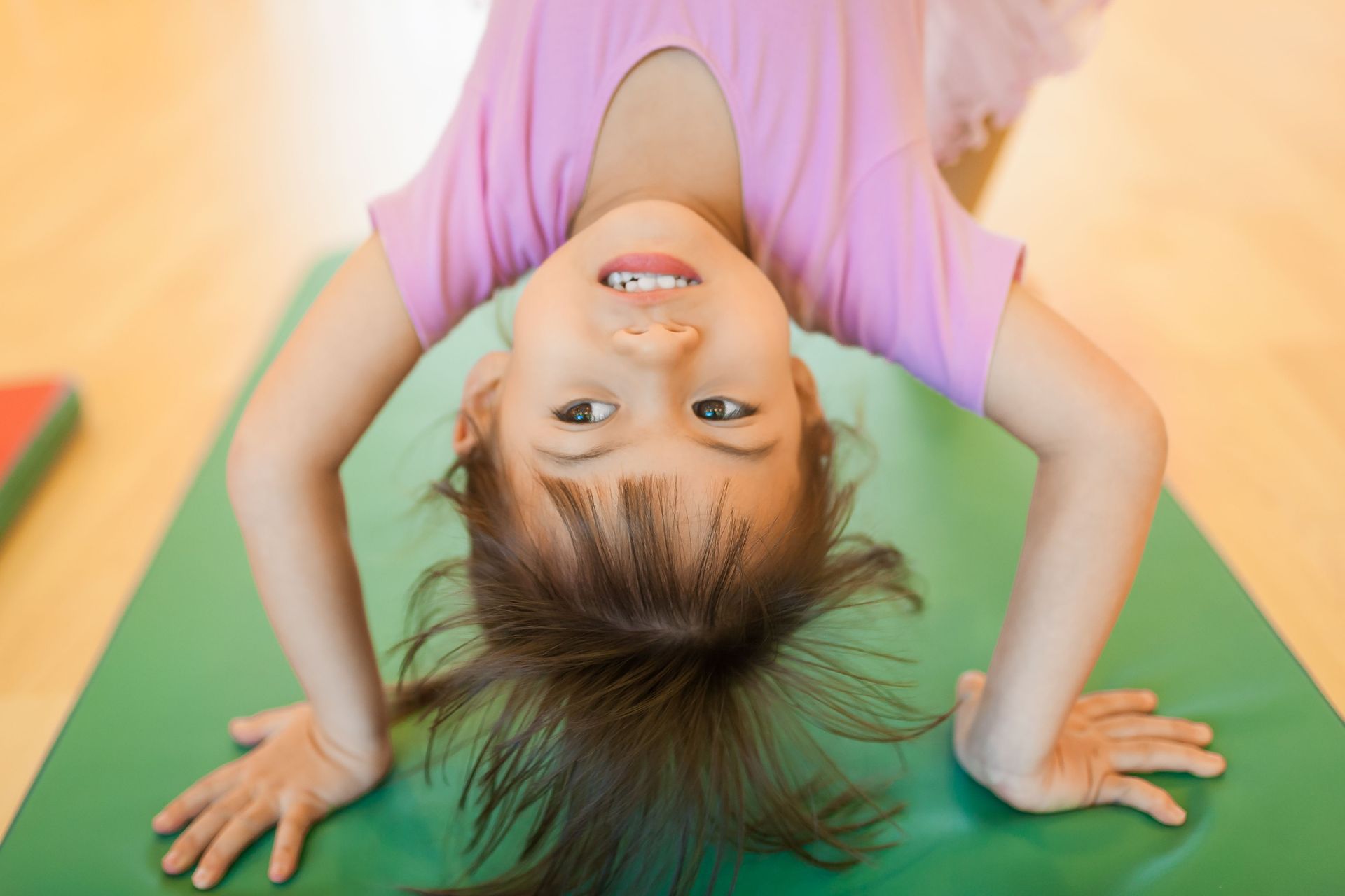 Child doing a handstand on a green mat, smiling in a purple shirt