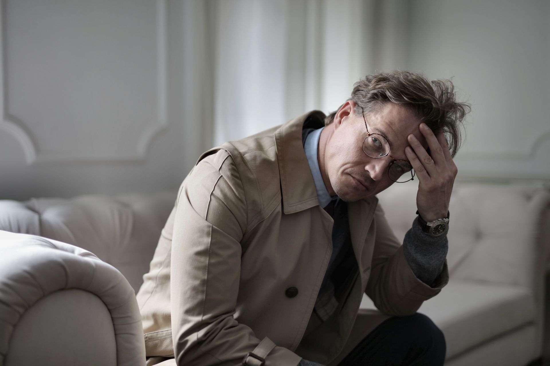 Distressed man seated on a sofa, holding his head in his hand in a dim living room.