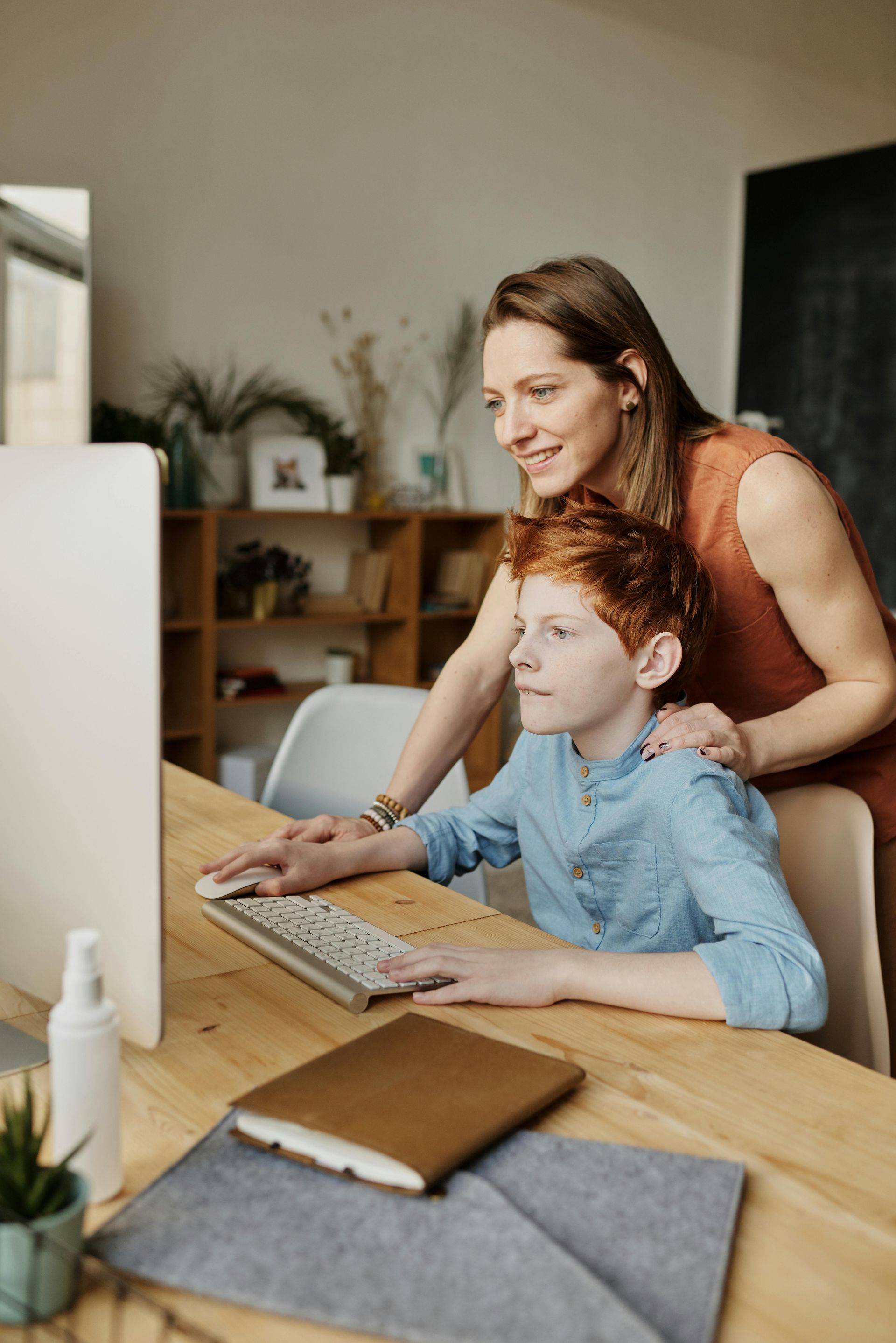 A woman is standing next to a boy using a computer.