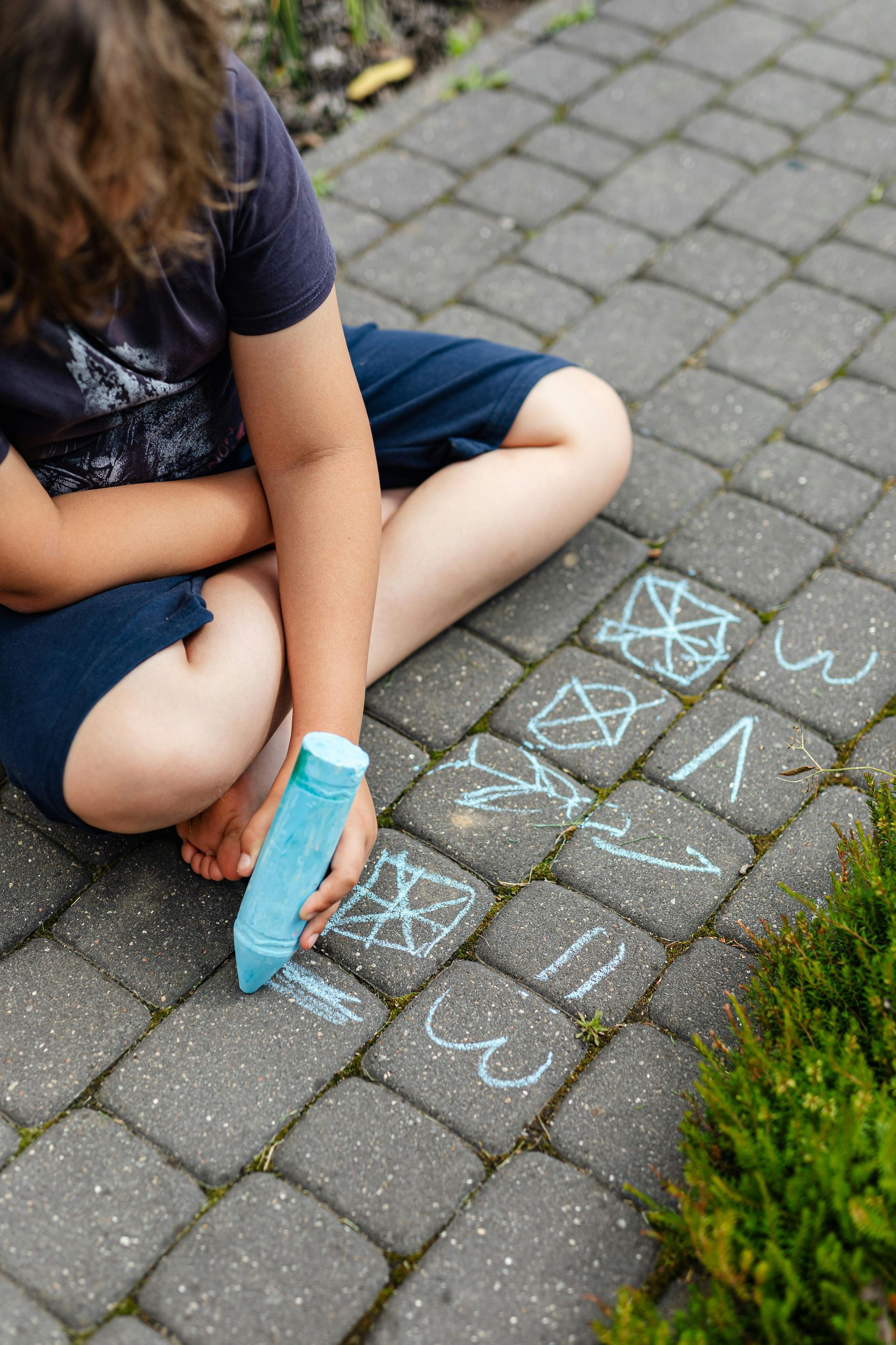 Child drawing with blue chalk on sidewalk hopscotch grid, with a foot in a blue cast nearby