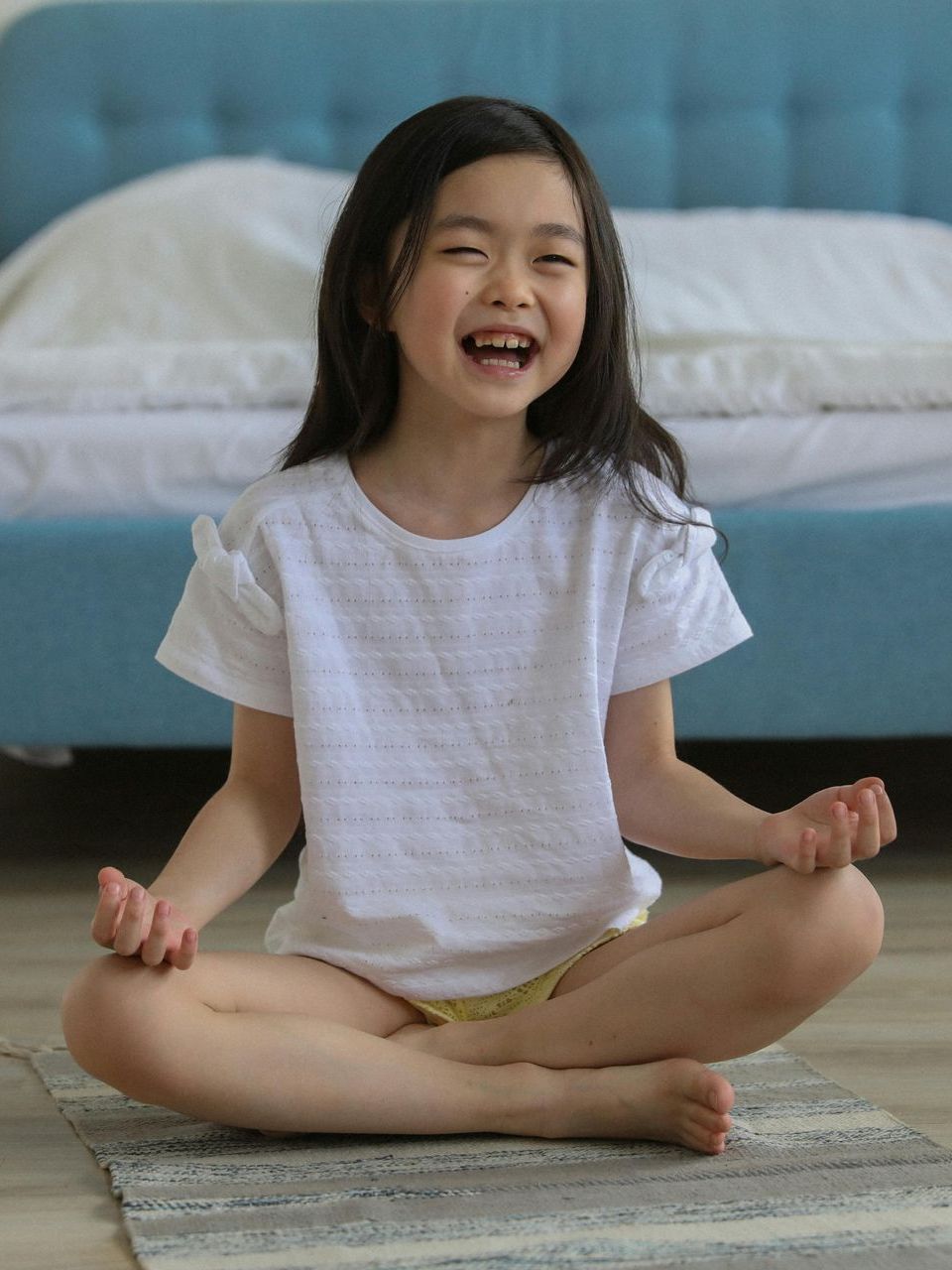 Young Asian girl sitting cross-legged, winking, smiling, in a white shirt and yellow shorts. Bedroom setting.