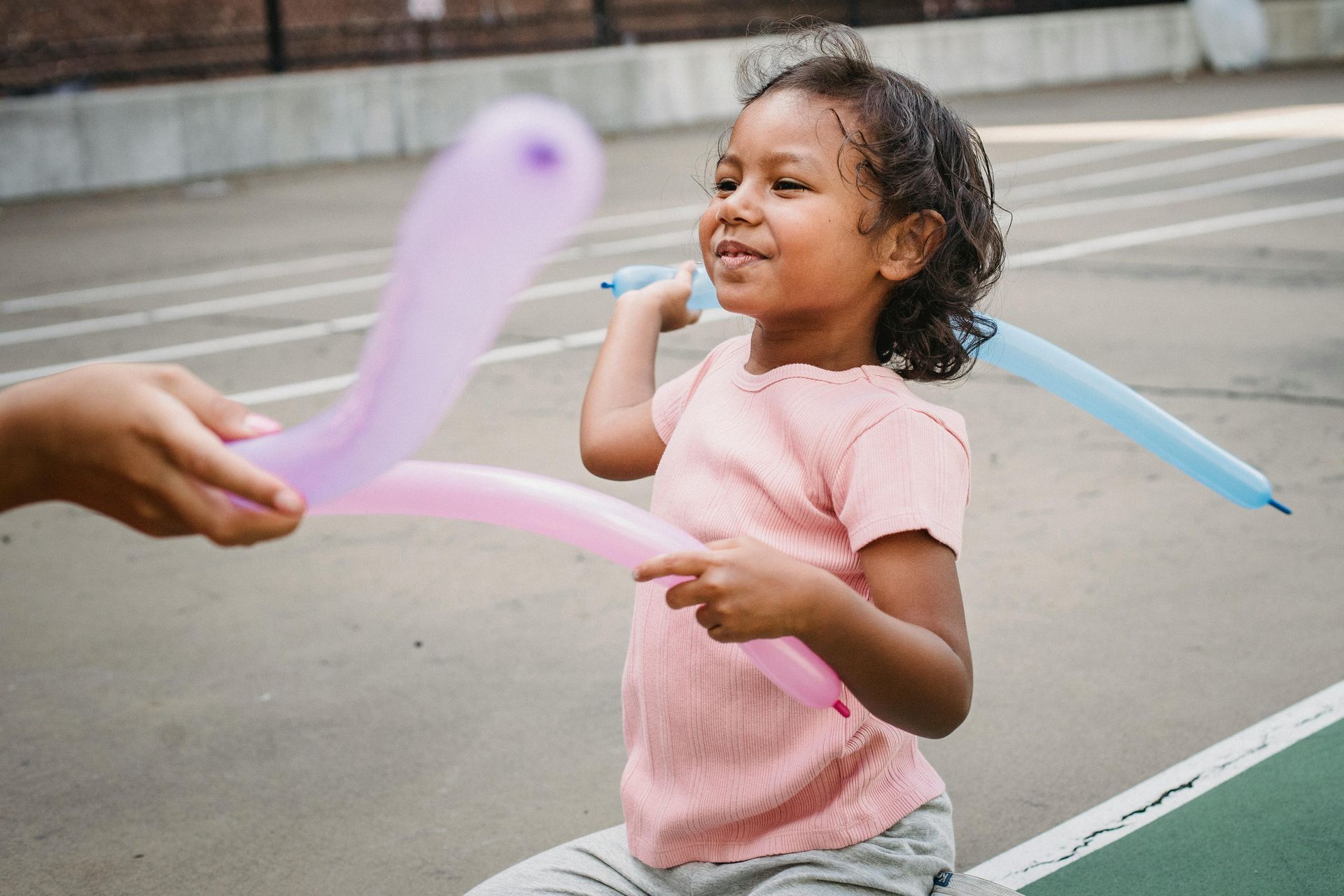 Child smiling while holding a blue balloon sword, reaching toward a pink balloon on a sidewalk.