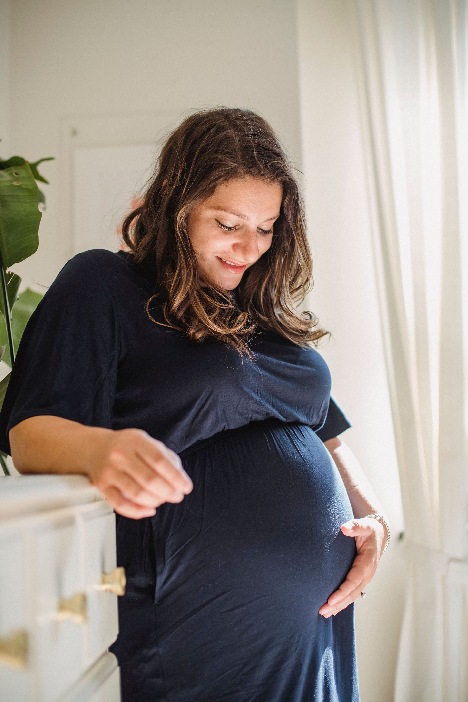 Pregnant woman in a navy dress standing by a window, looking down and gently touching her belly