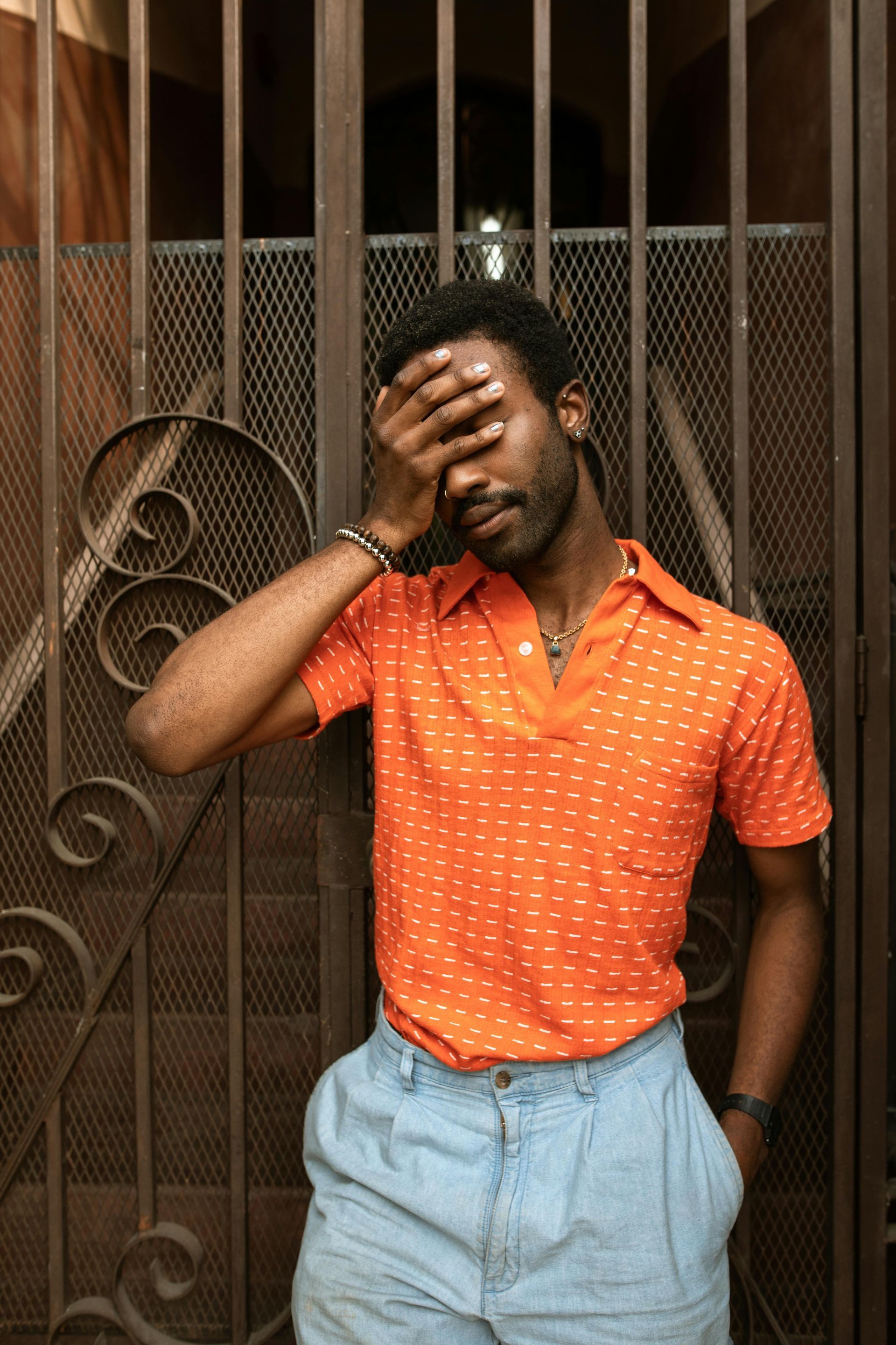 A man in an orange shirt and blue shorts is covering his face with his hand.