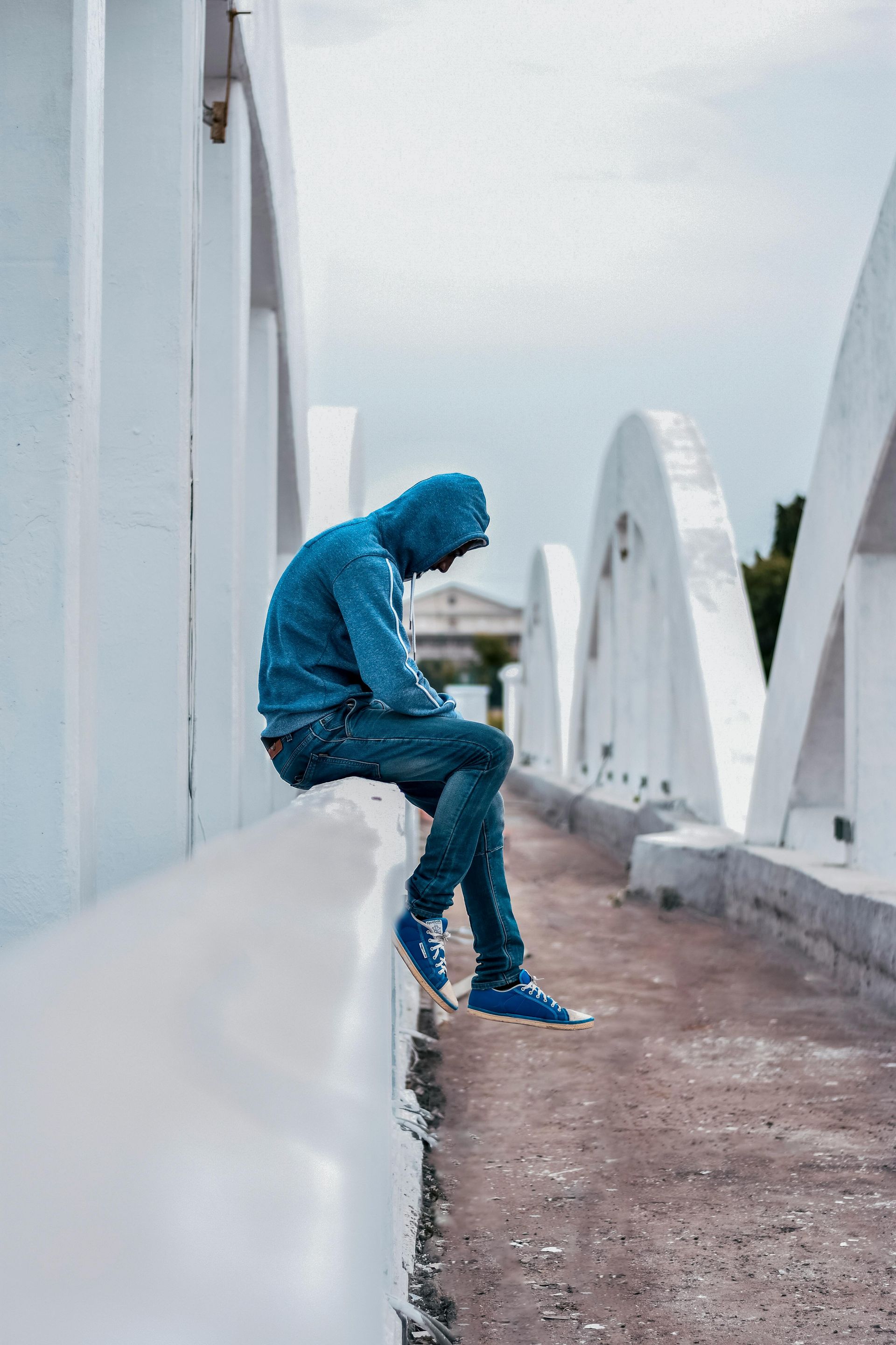 Person in a blue hooded jacket sits on a bridge railing in a misty outdoor setting.