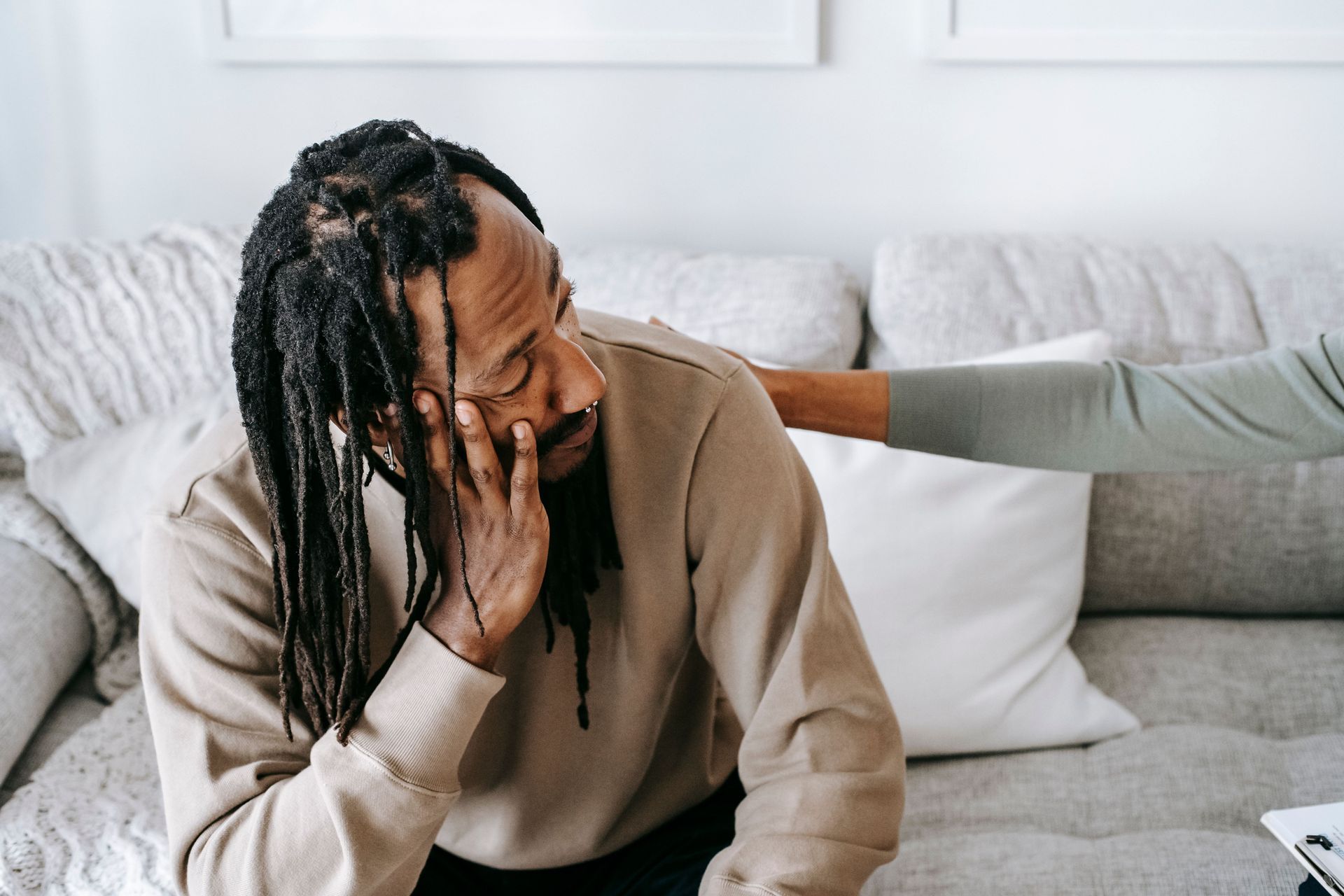 Person sitting on a couch, holding their face while another person comforts them with a hand on the shoulder.