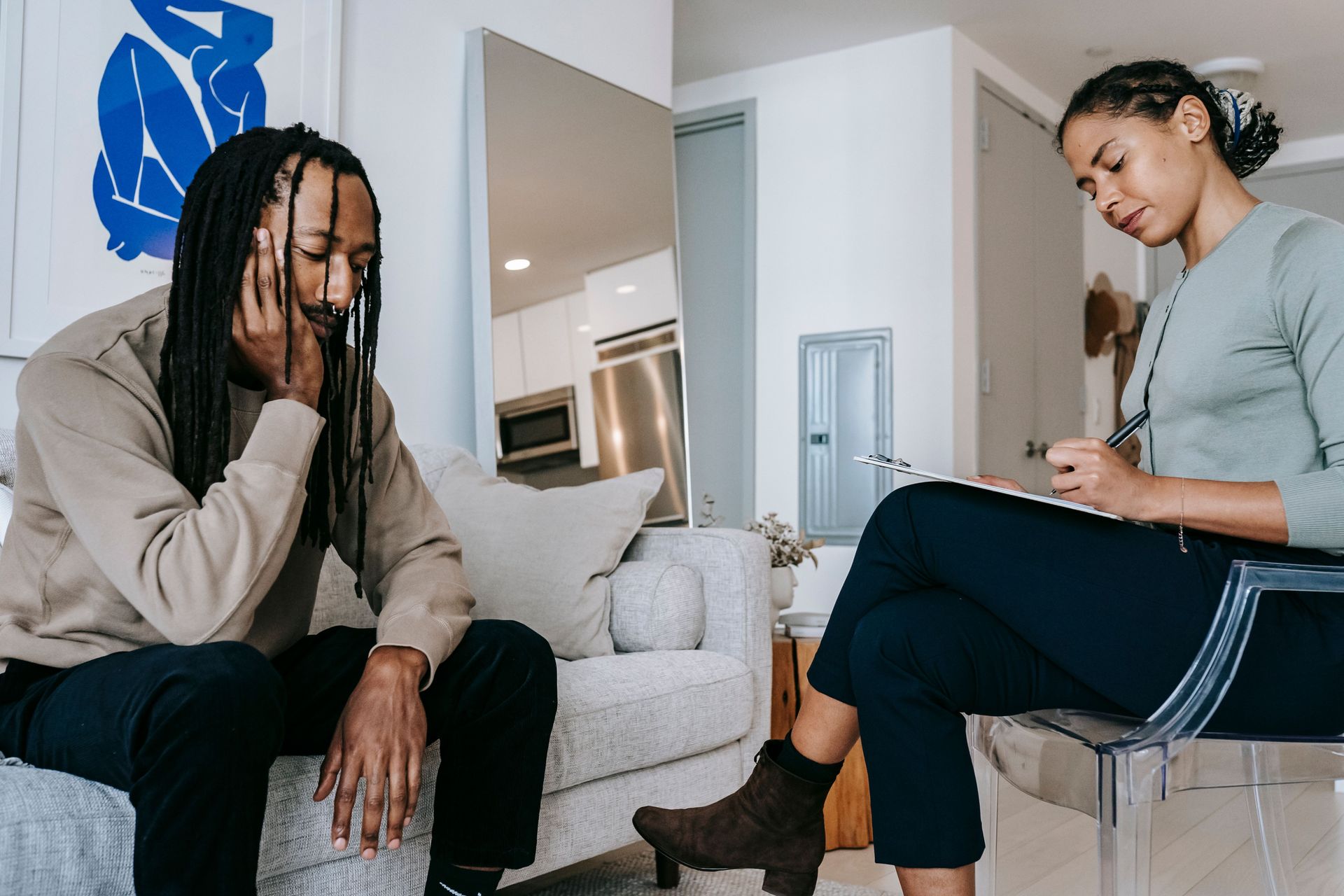 Two people sit talking in a bright offic, one holding a notebook and the other listening on a couch.