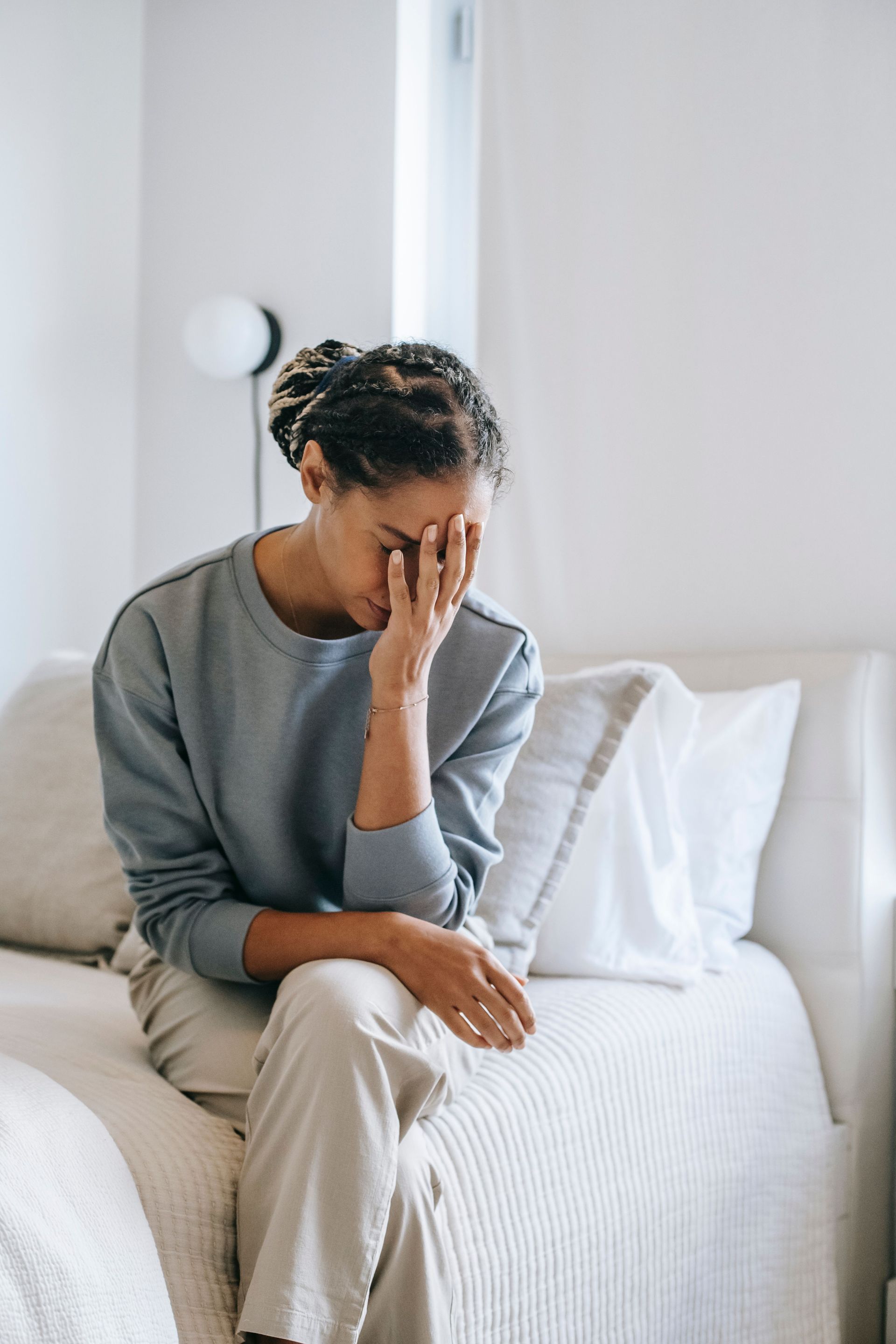 Person sitting on a bed with head in hand, appearing stressed in a bright bedroom