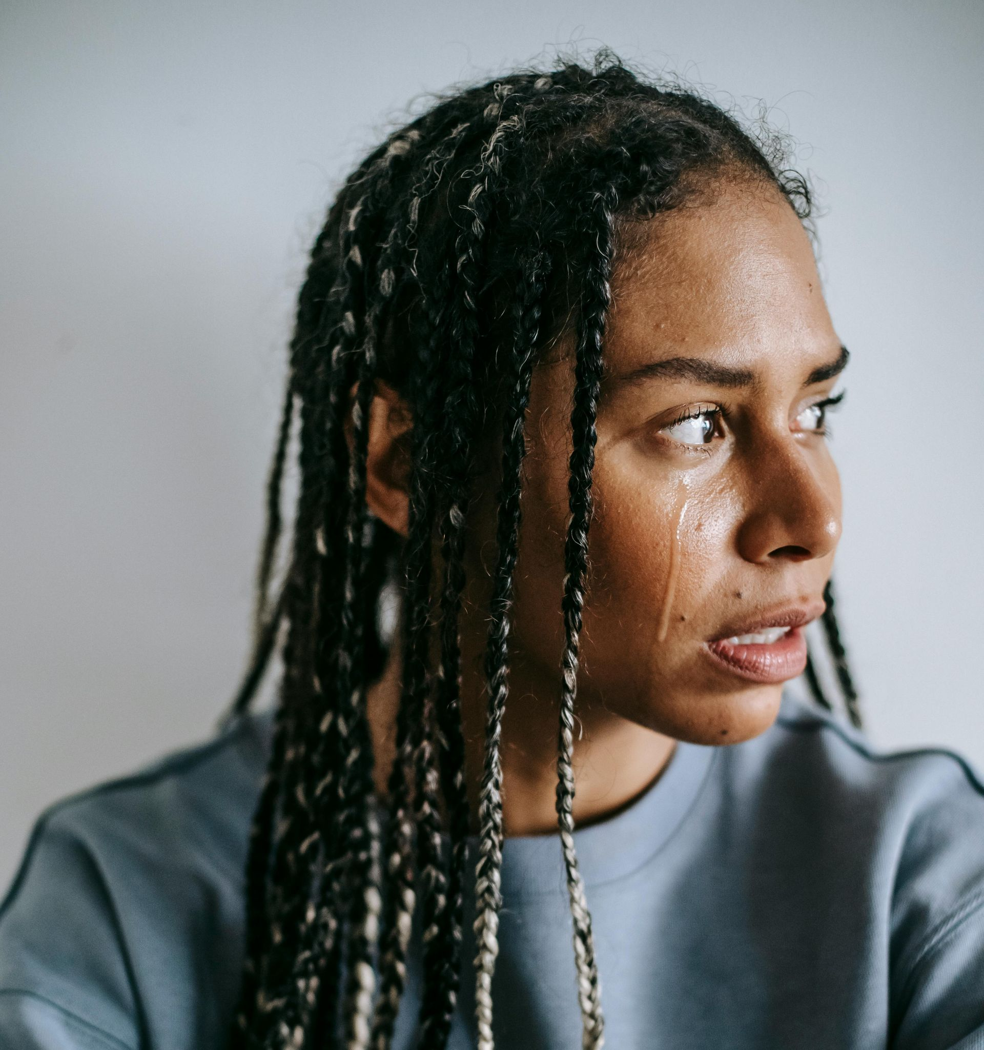 A person with braided hair and a tear-streaked, distressed expression looking to the side while wearing a blue sweatshirt.