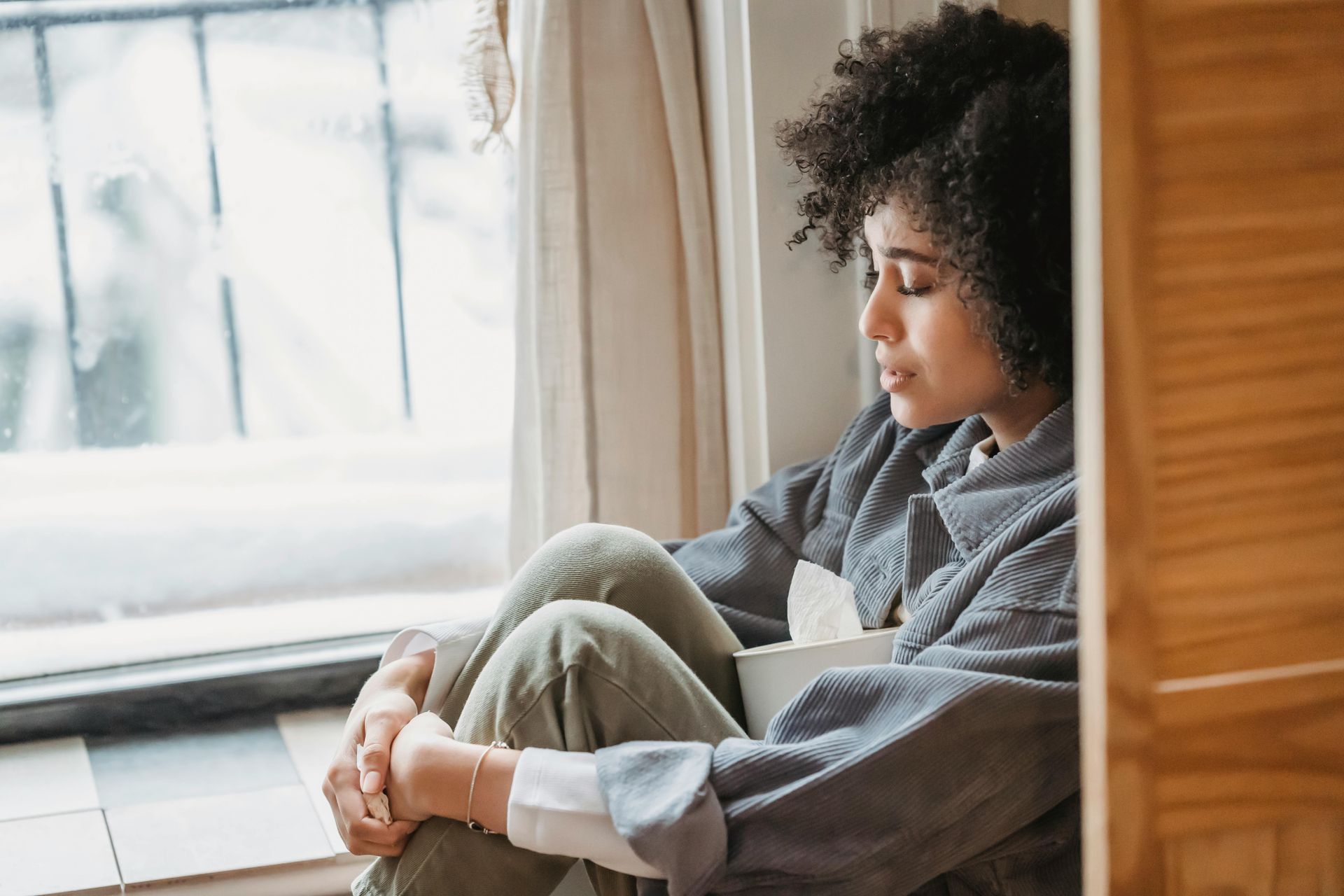 Person sitting on the floor, crying by a window, looking outside with knees drawn up.