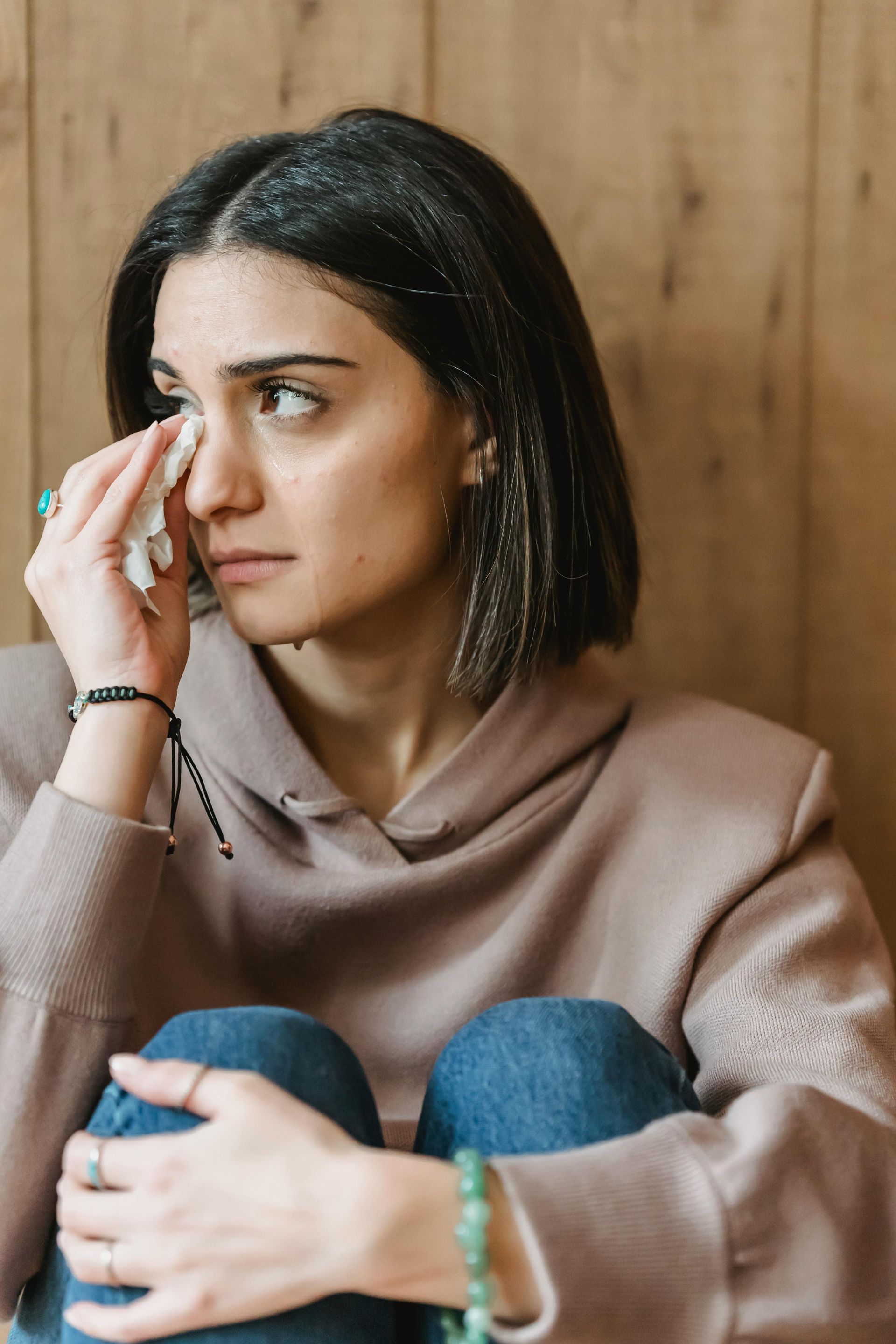 Woman in a pink hoodie sitting against a wooden wall, looking to the side and crying.