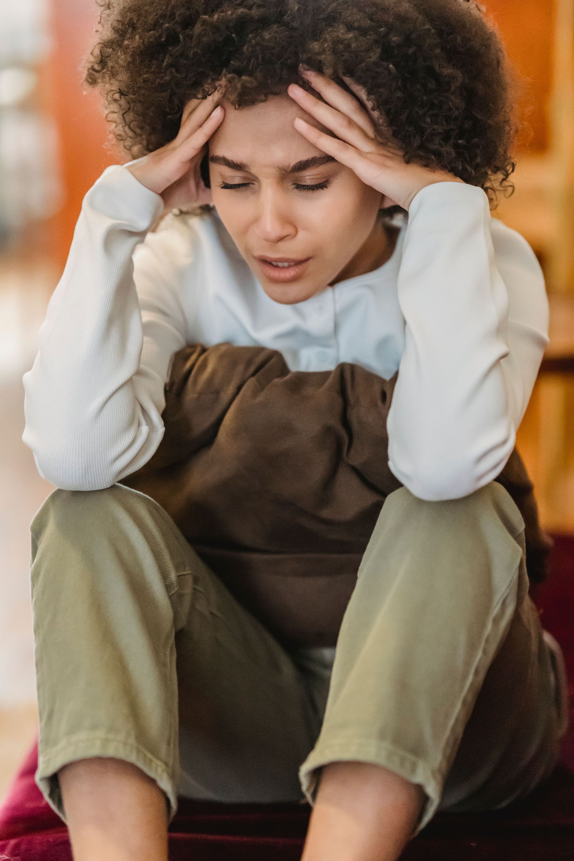 Person sitting on the floor with head in hands, looking distressed, wearing a white sweater and olive pants.
