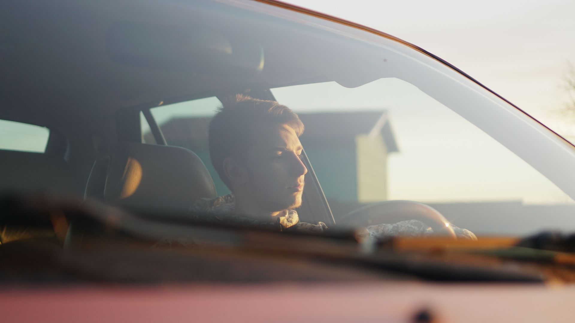 Person driving a car at sunset, seen through the side window with warm light on their face.