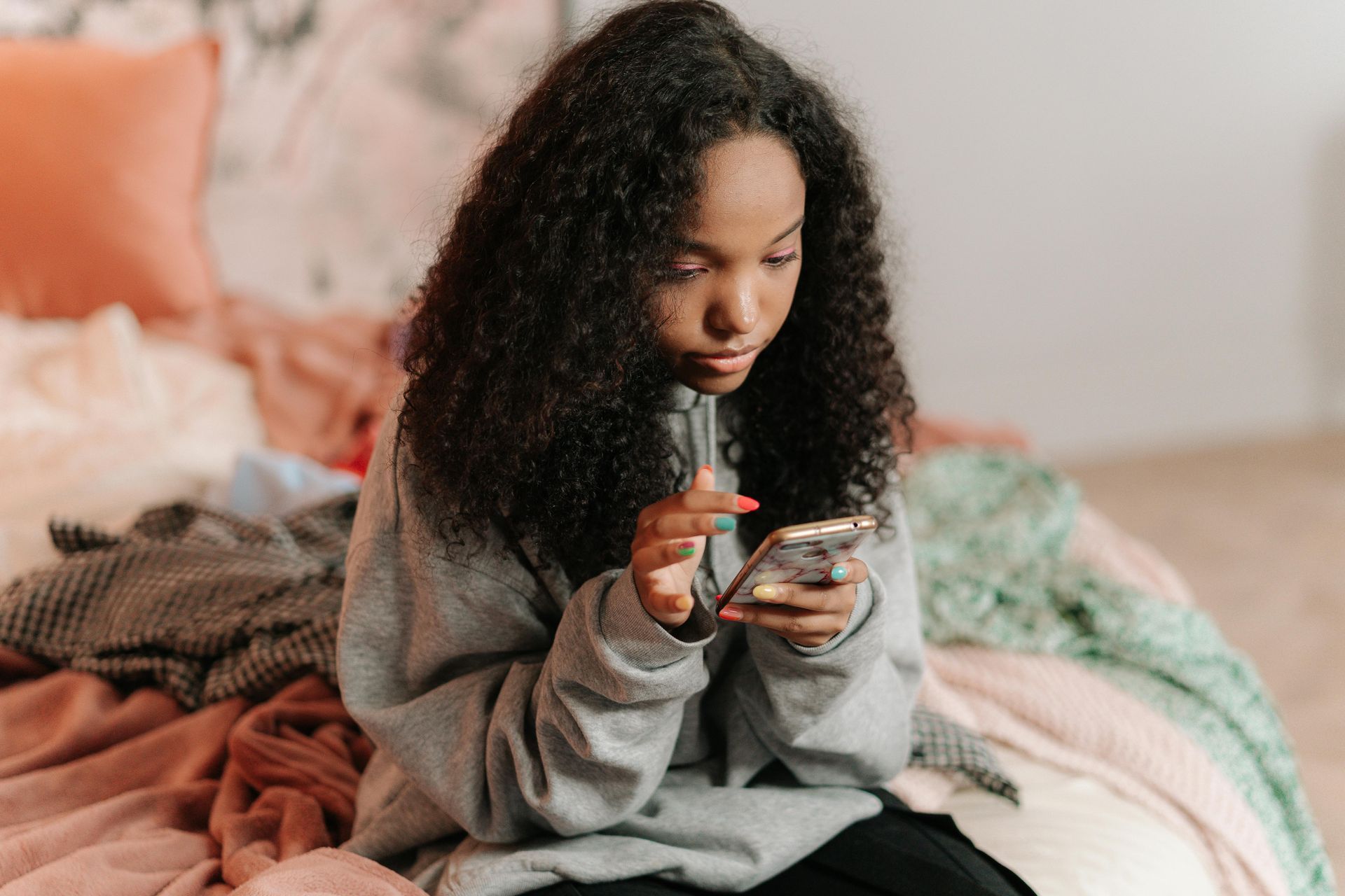 A young Black woman in a grey sweatshirt uses her phone, sitting on a bed with colorful blankets.