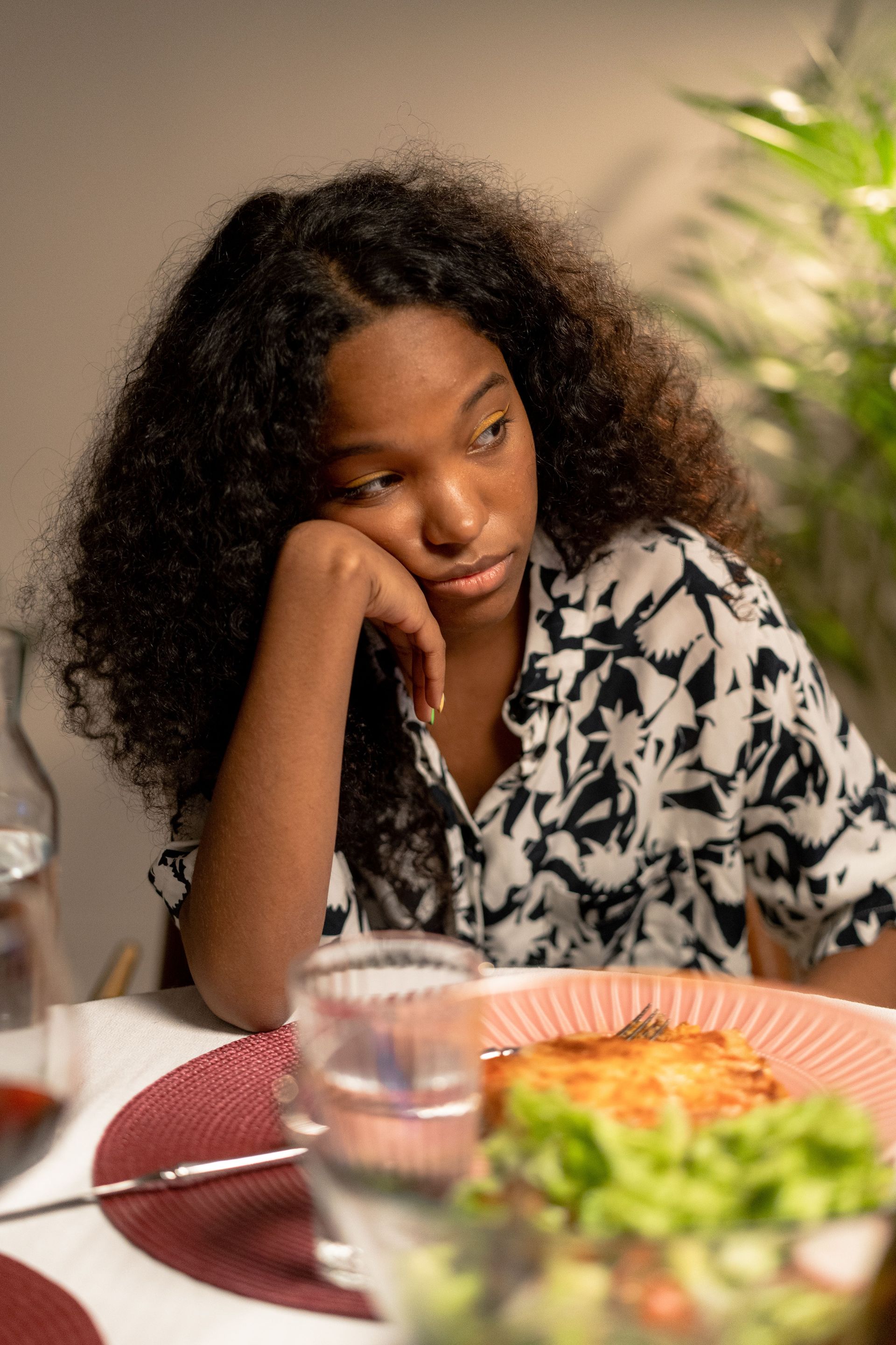 Teen resting their head on their hand at a table with a salad and pizza, looking bored.