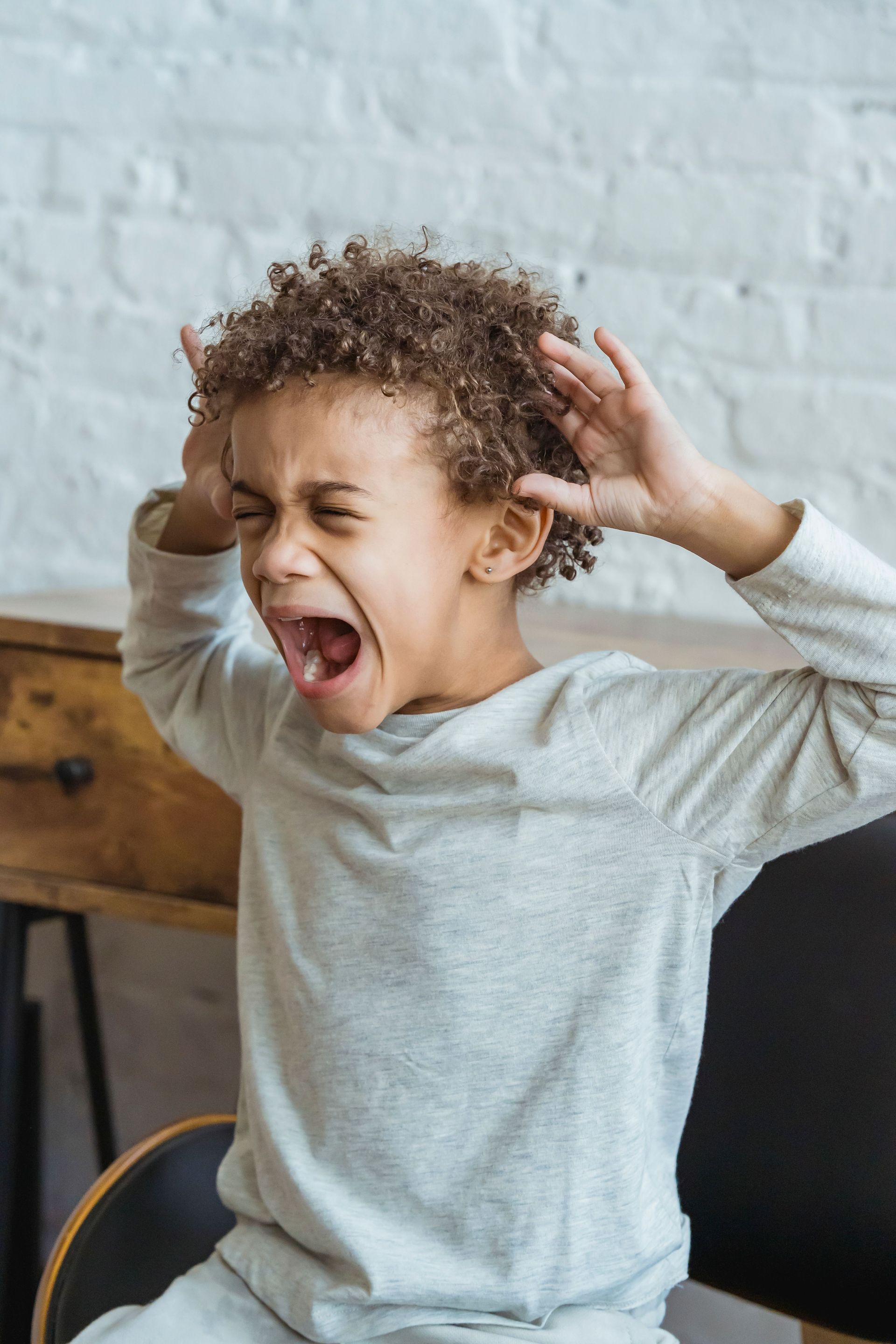Child in a gray sweater yelling with hands raised near ears, seated indoors against a white brick wall.