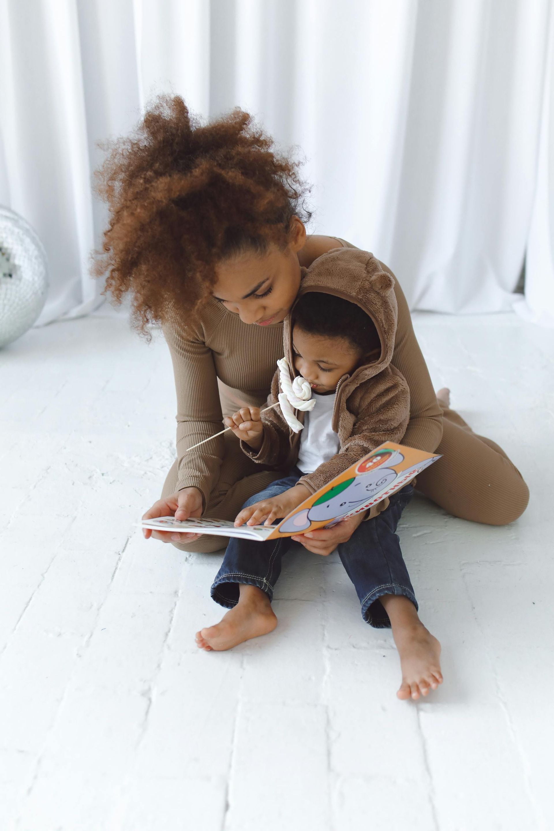 Woman and child reading a book together on a white floor. Woman in beige outfit; child in hoodie.