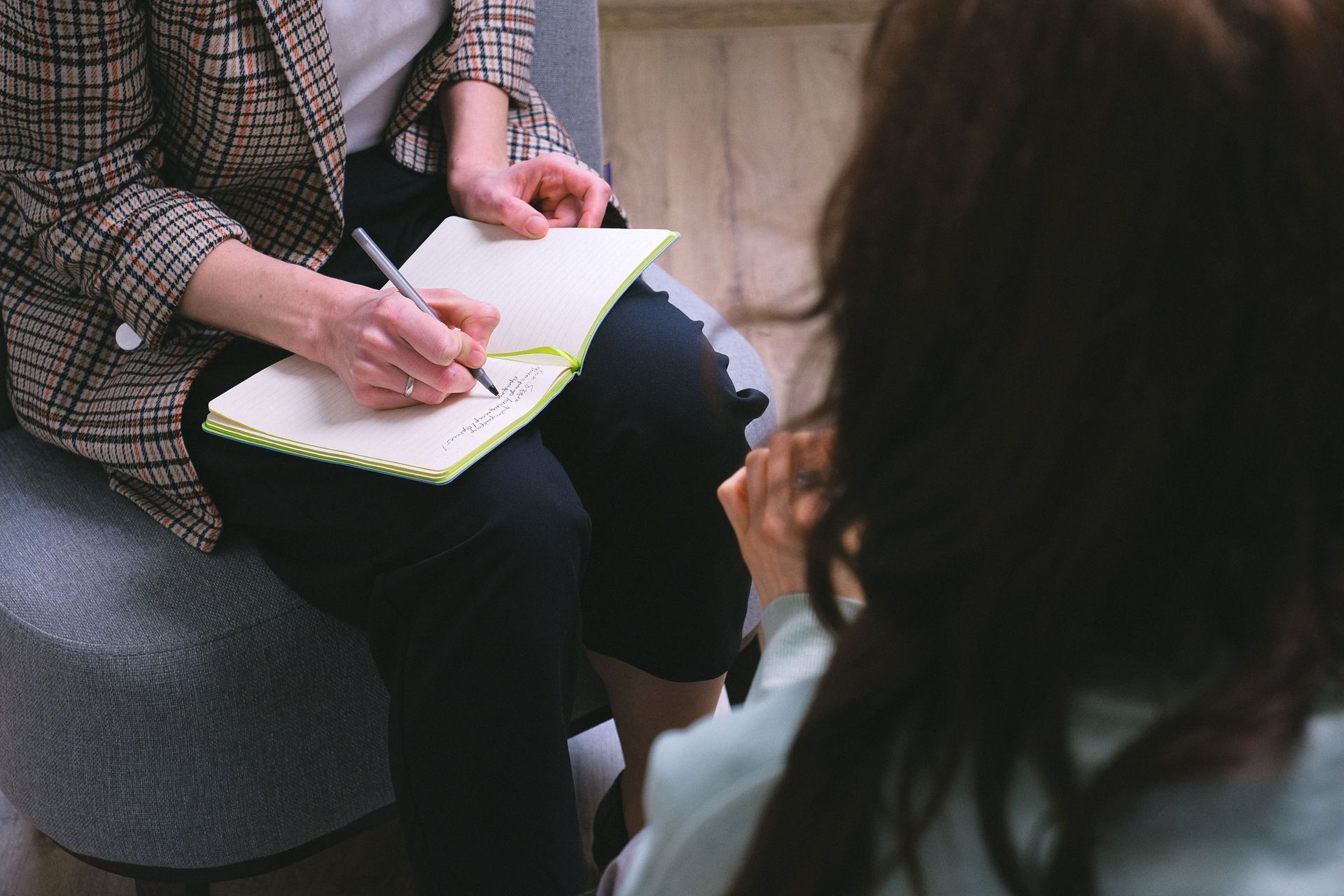 Person writing in a notebook during a seated conversation, with another person blurred in the foreground.
