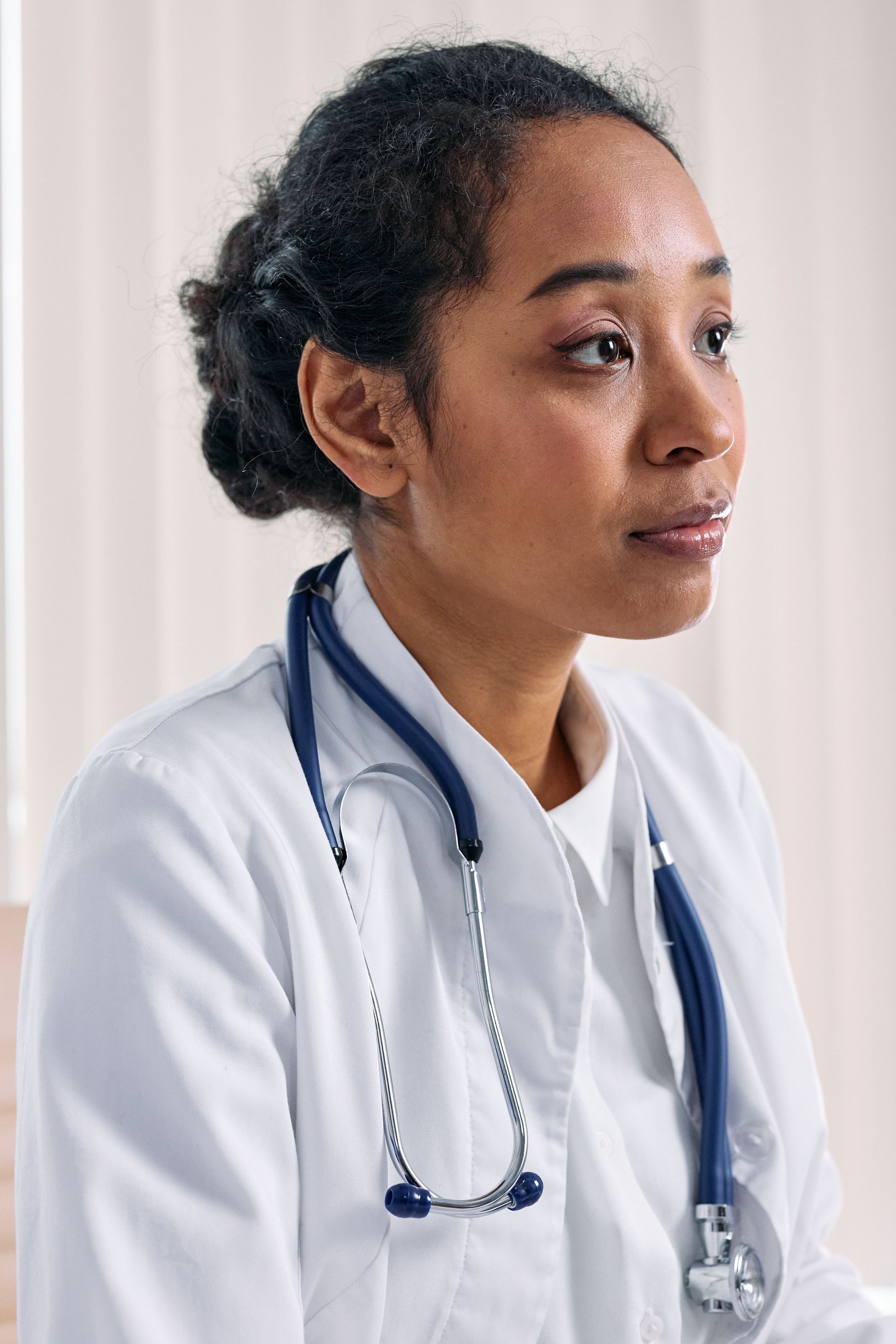 Doctor in a white coat with a stethoscope, looking to the side in a bright clinic.