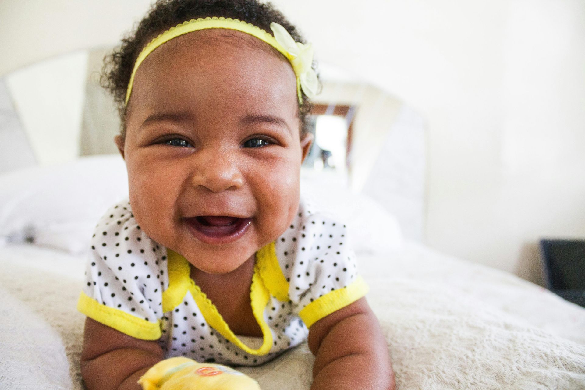 Smiling baby in polka dot outfit, yellow headband, on a white bed.