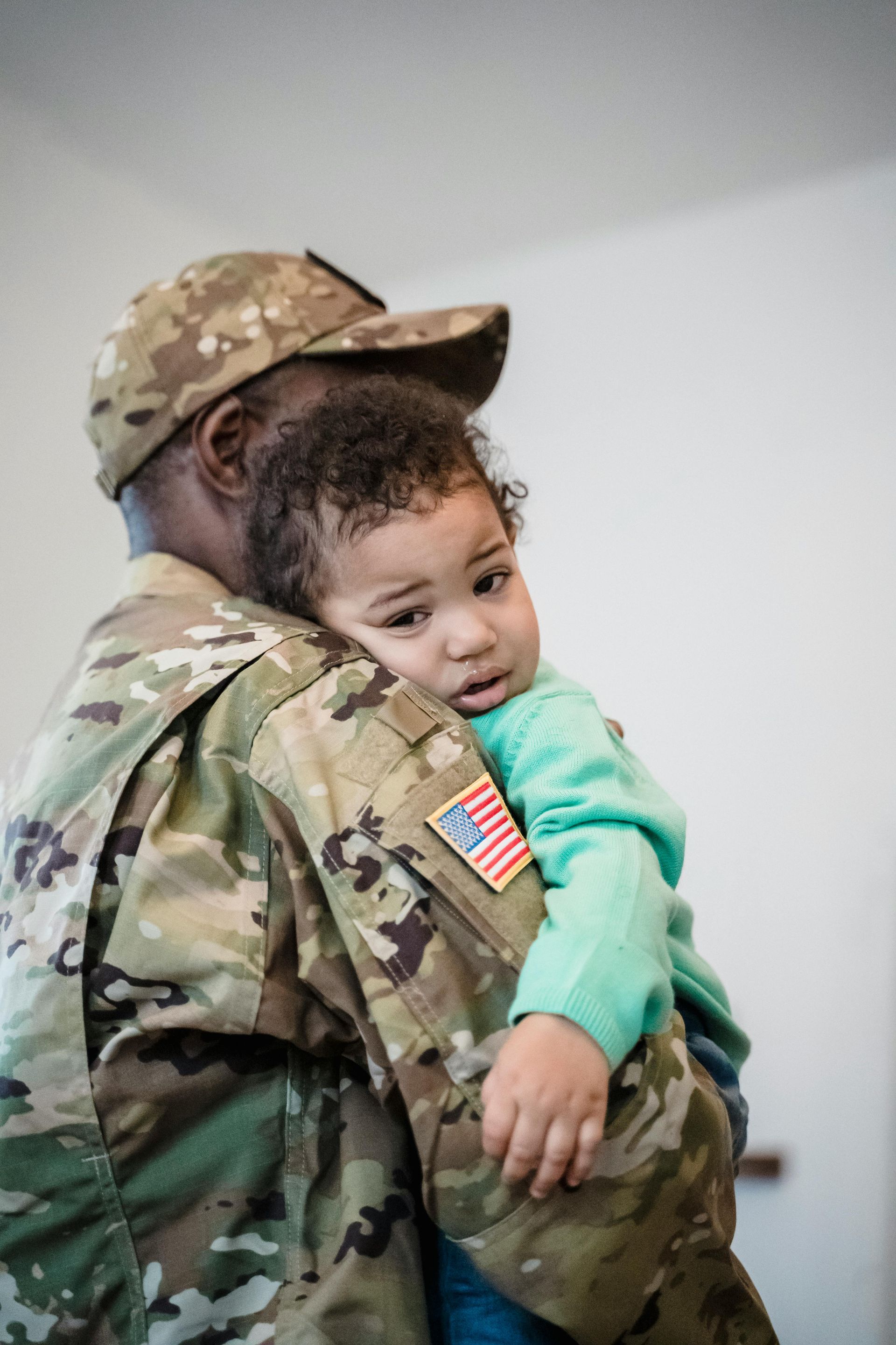 Soldier in camouflage holds a child, indoors. The child looks at the camera.