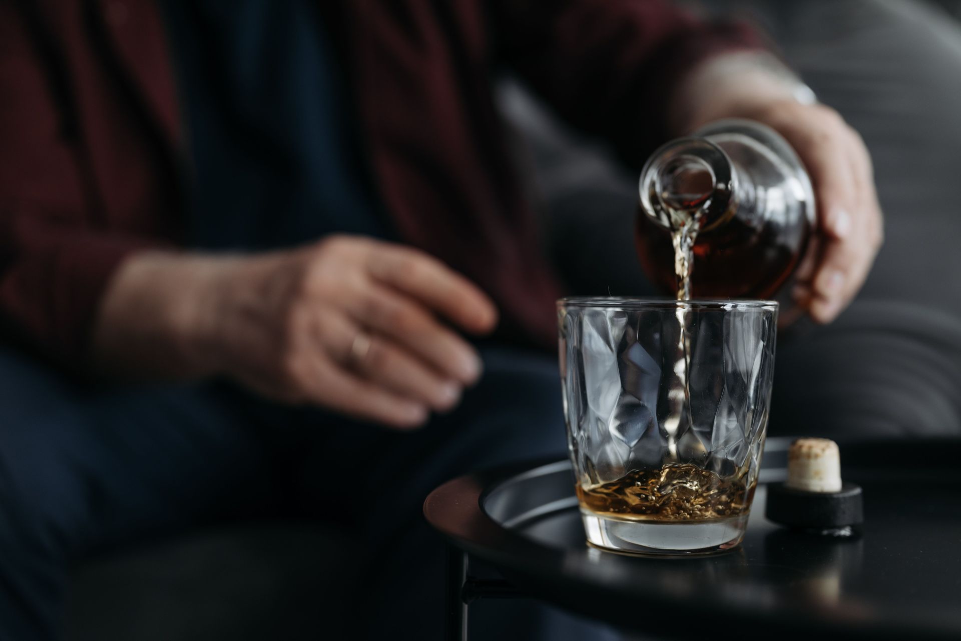 Whiskey being poured into a glass beside a seated person in a dimly lit setting