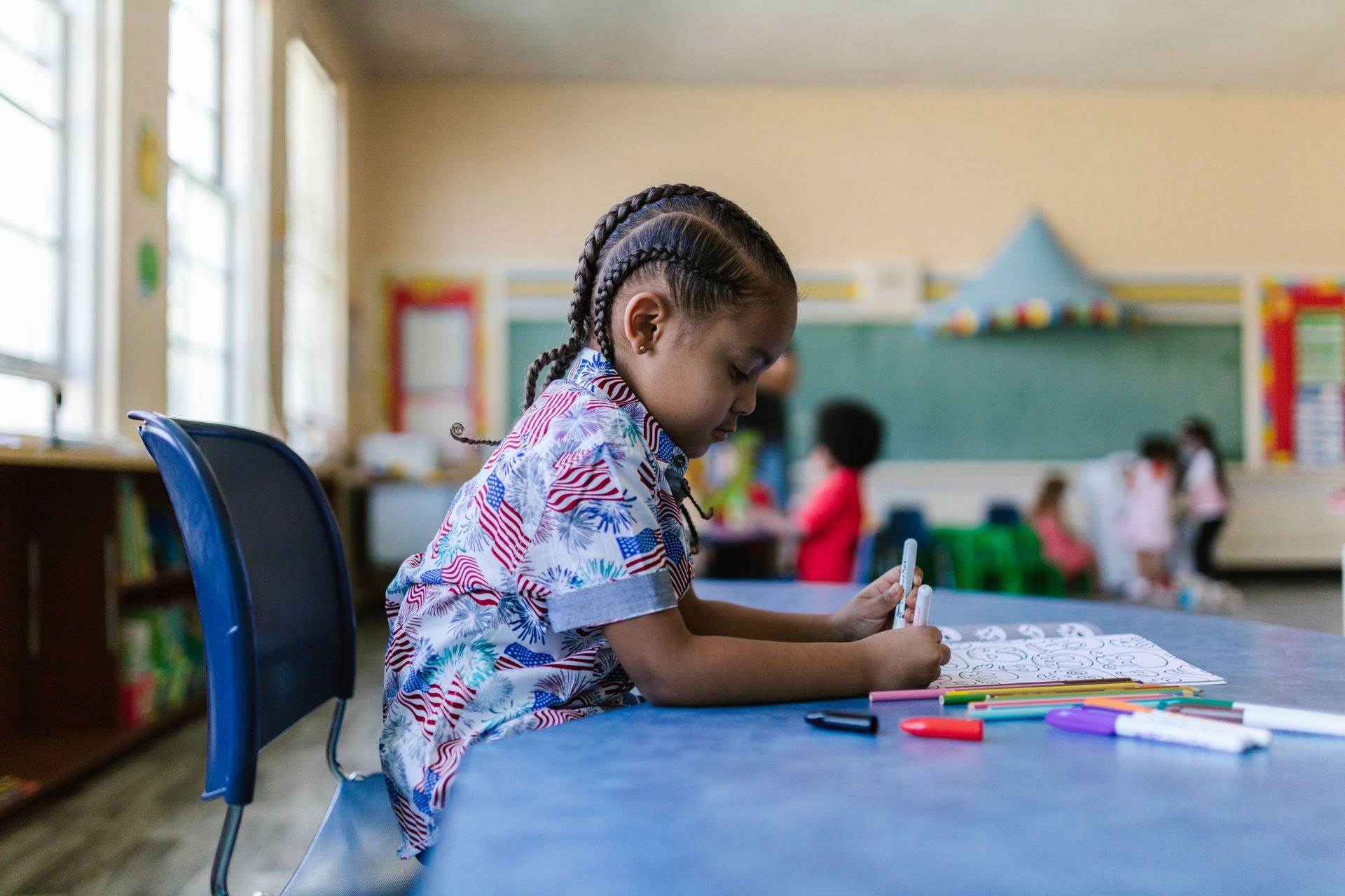 Child drawing at a table in a bright classroom, with colored markers and papers nearby.