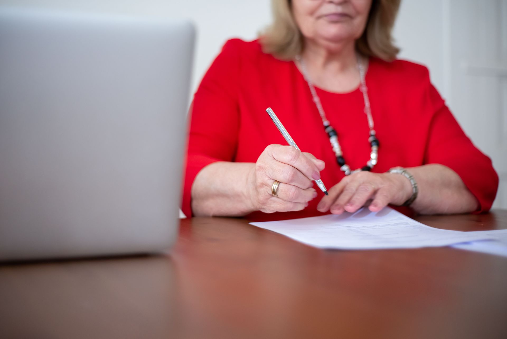 Person in red shirt writing on paper beside a laptop at a desk